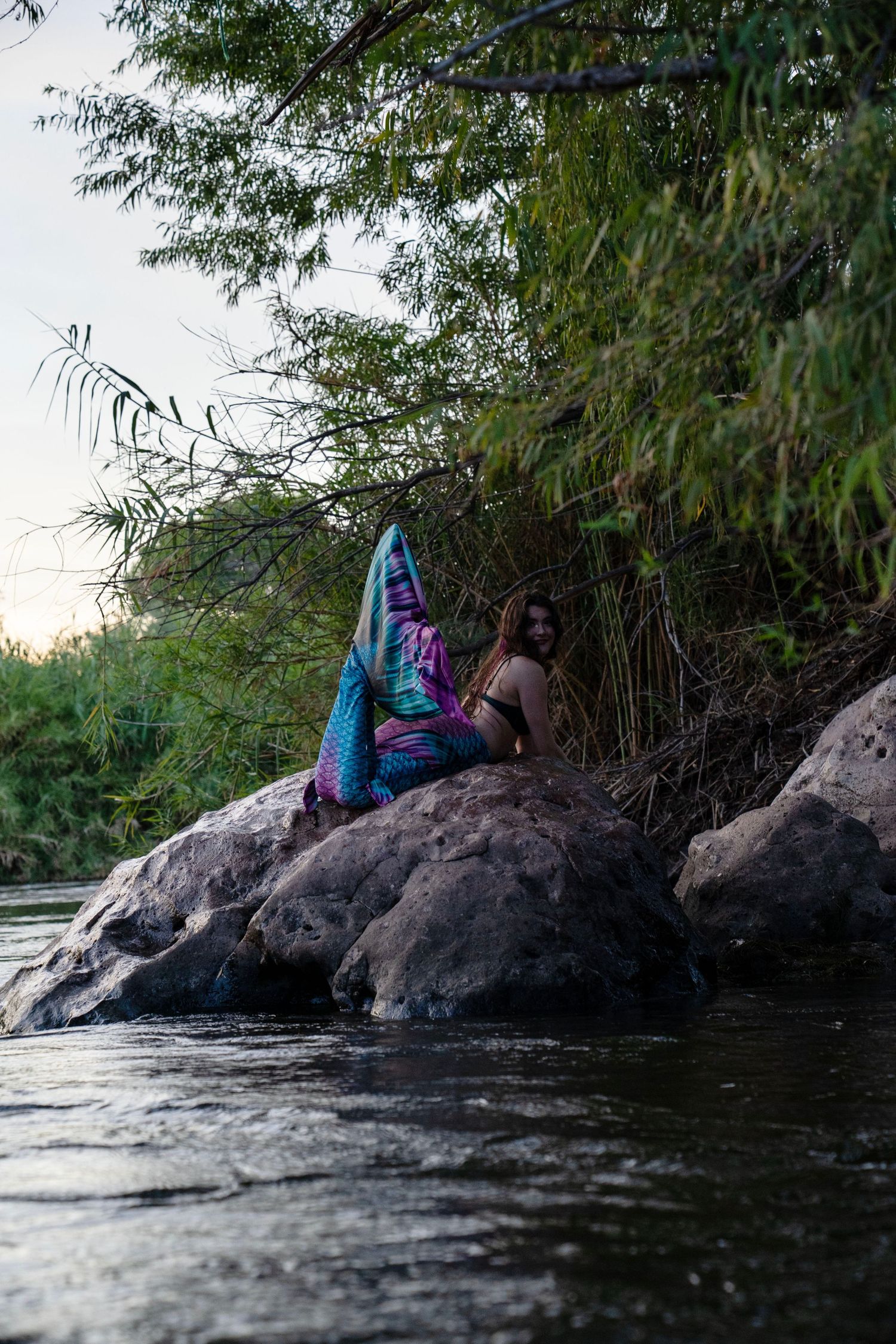 A woman in a colorful mermaid costume sits on rocks beside a calm river with dense green trees in background. Fantasy Photoshoot
