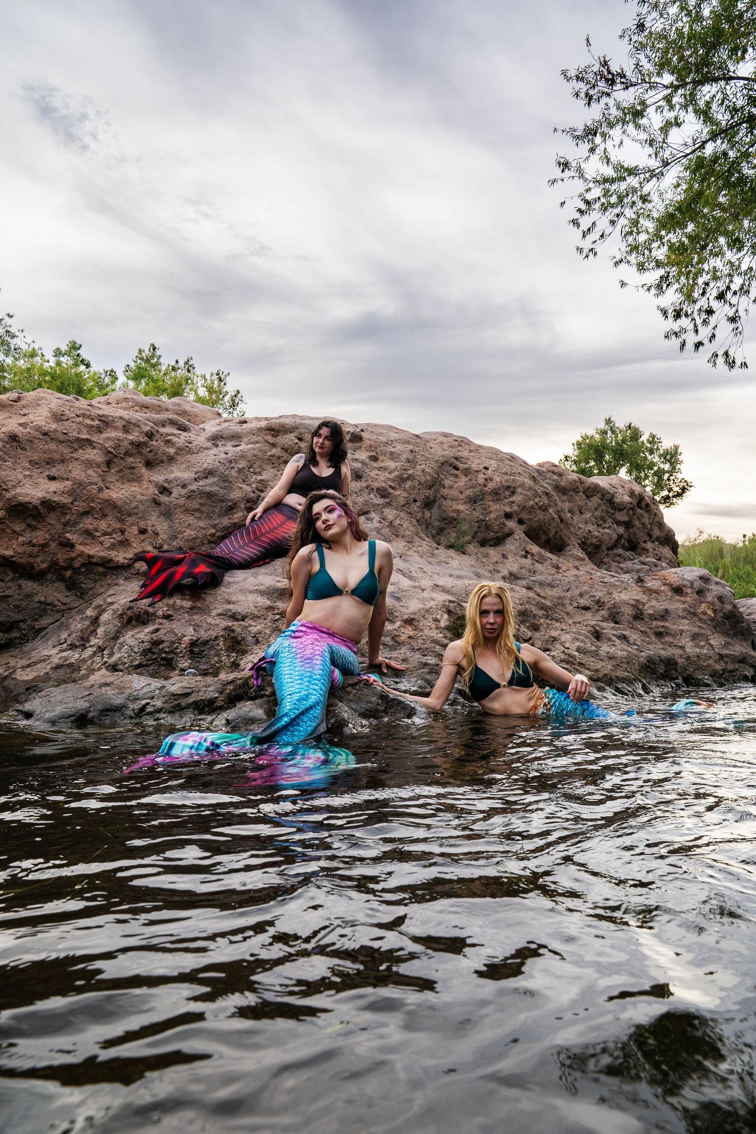 Three women in colorful mermaid costumes pose on rocks and in water at a scenic outdoor location. Fantasy Photoshoot