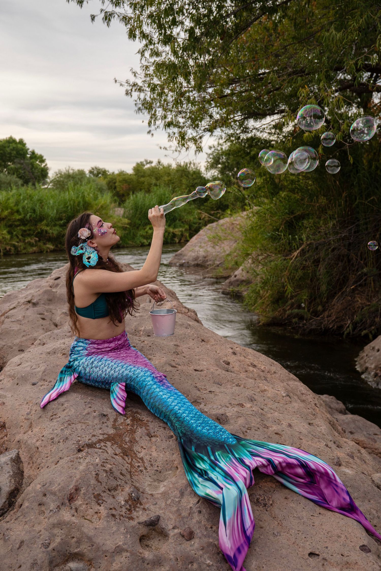 A woman in a colorful mermaid costume blows bubbles while sitting on rocks by a river. Fantasy Photoshoot