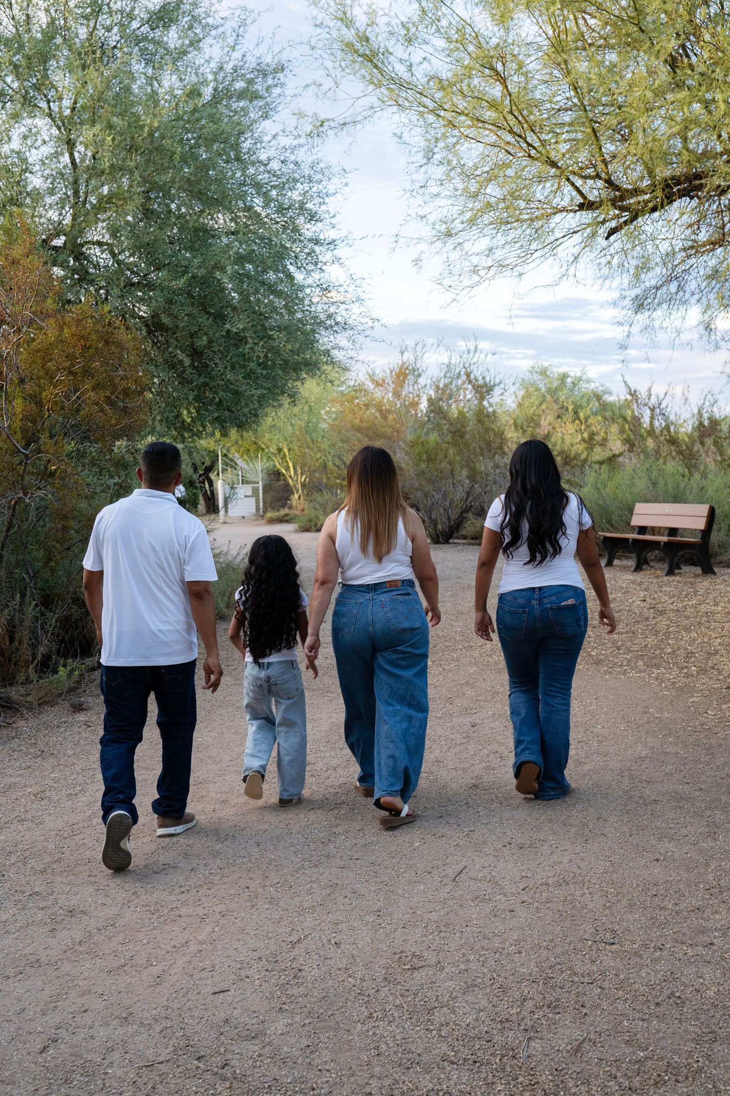 Family of four walking away on a desert trail, wearing white shirts and jeans, surrounded by green trees.