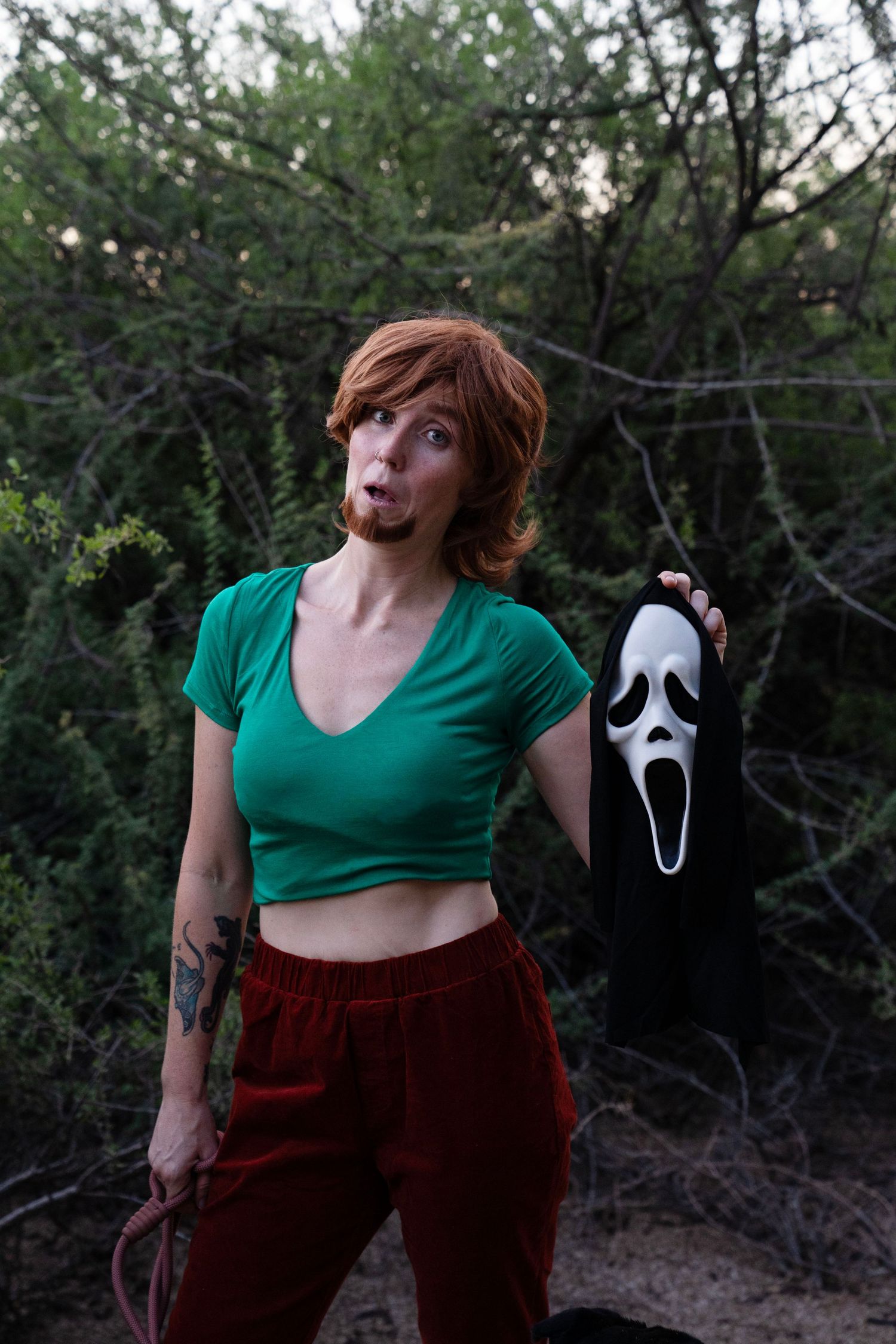 Woman in a green crop top and red velvet pants poses outdoors among bushes, holding a Ghostface-style mask in a Halloween cosplay.