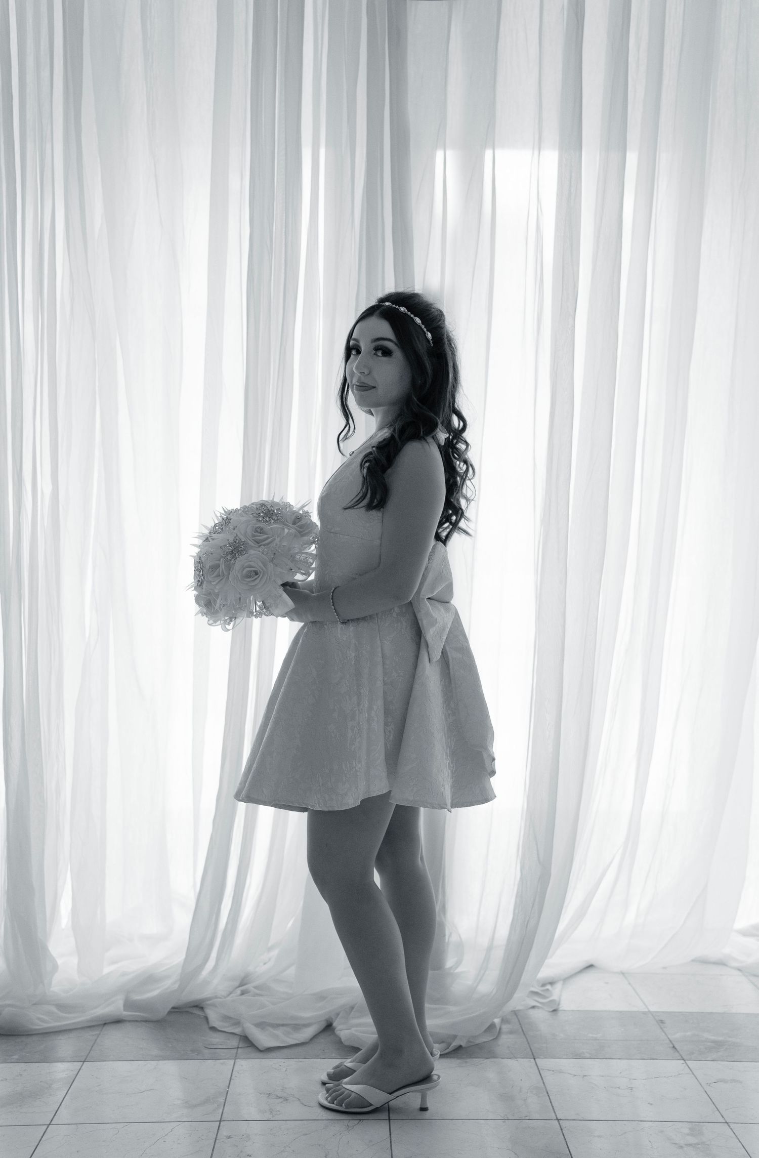 Black and white photo of a young woman in a short dress holding a bouquet, posing elegantly in front of white curtains.