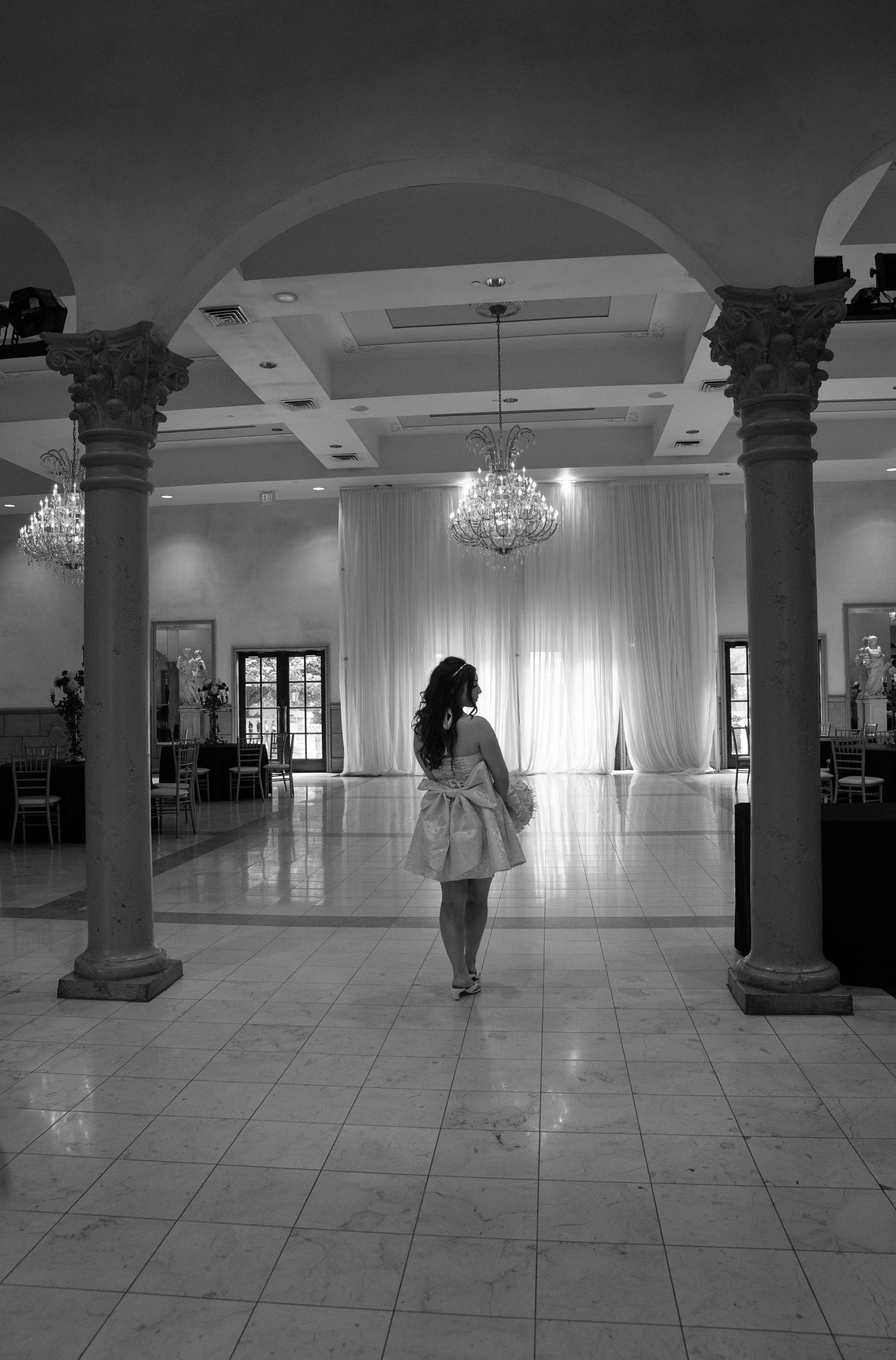 Black and white image of woman in pink dress standing in elegant ballroom with chandeliers.