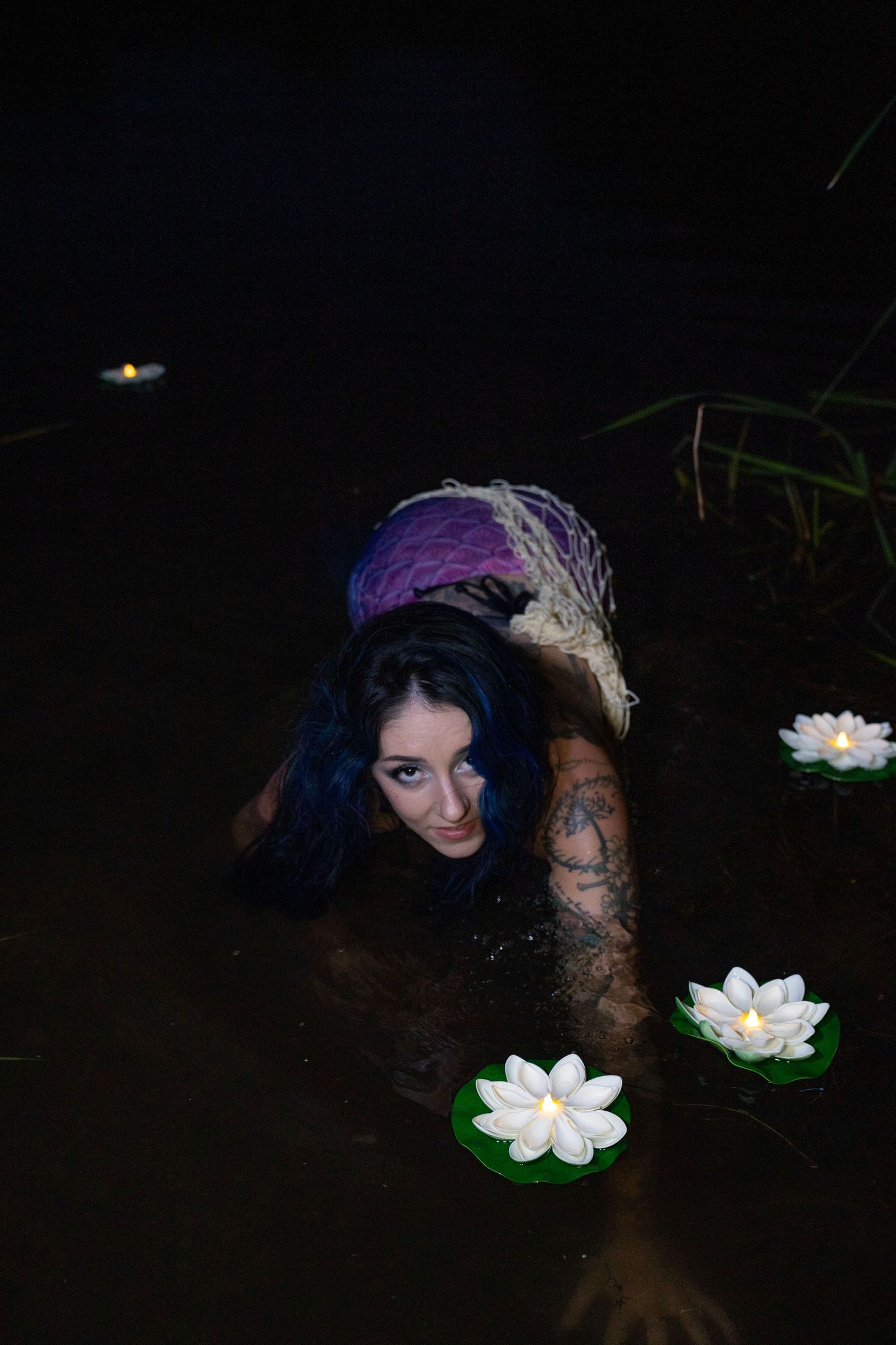 Tattooed woman in mermaid tail submerged in dark water surrounded by floating white lotus flowers.