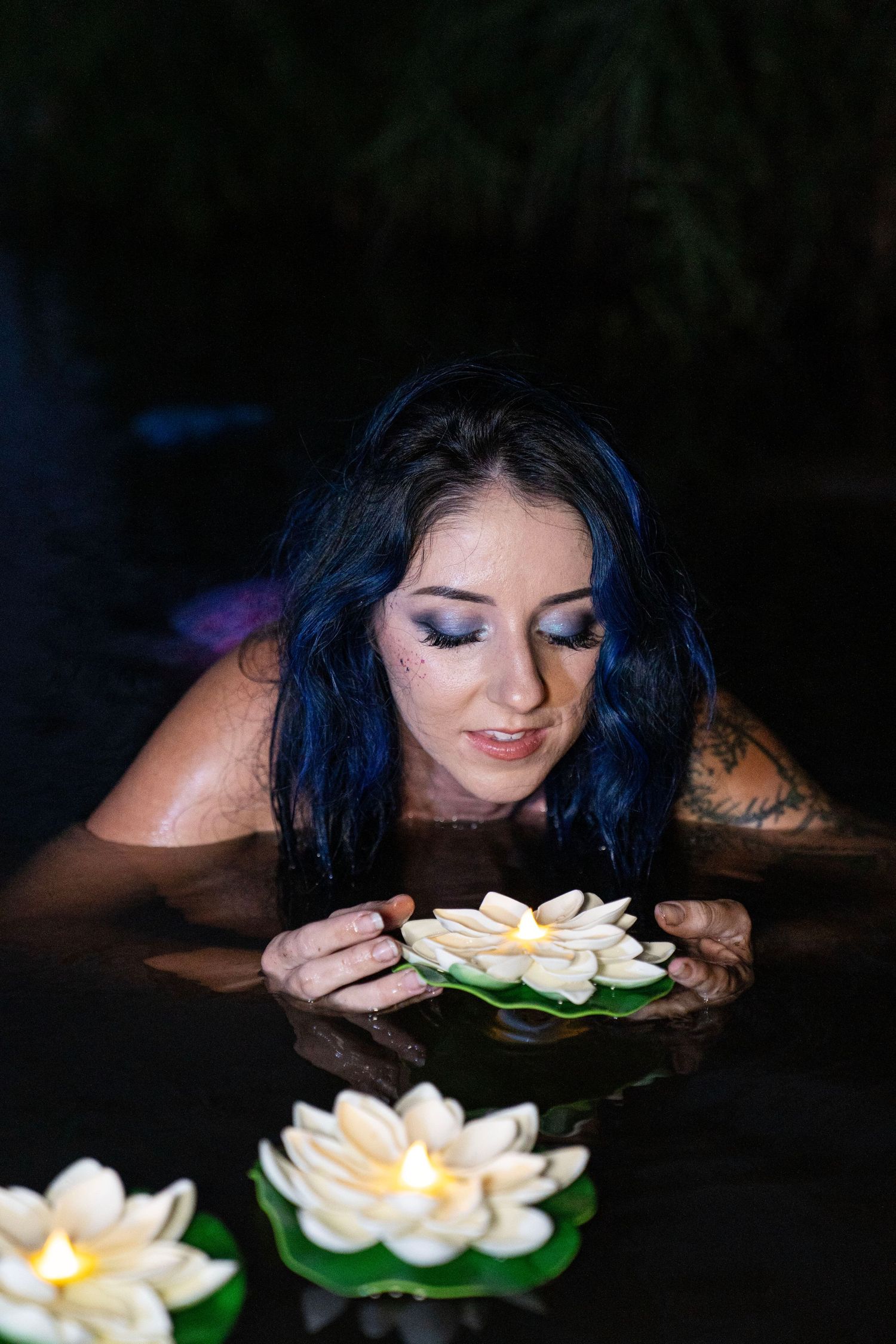 Blue-haired woman with eye makeup smelling floating lotus candles in dark water.