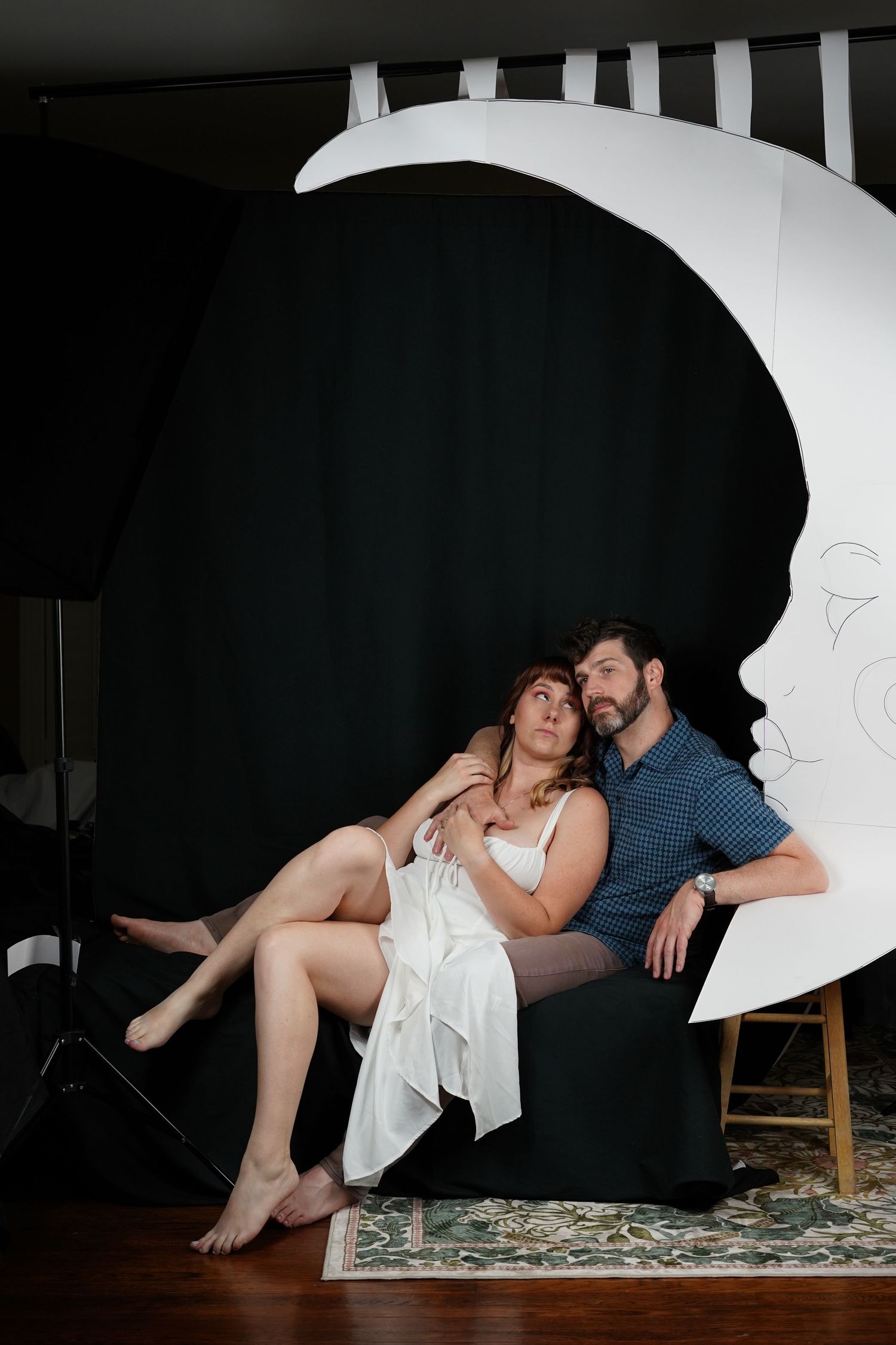 A couple sits together on a chair in front of a large decorative paper moon backdrop against a black curtain.