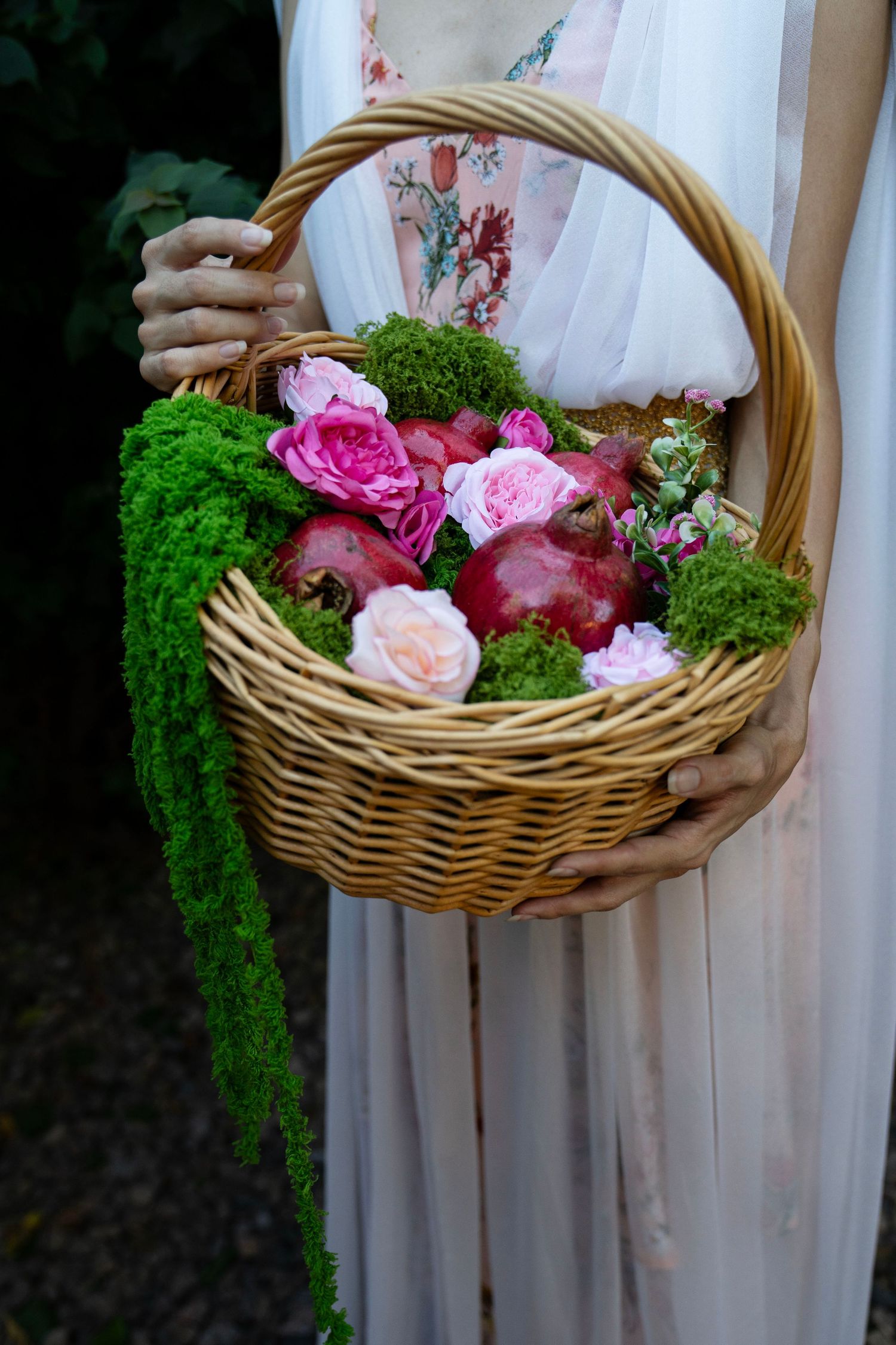 Woman in a flowing white dress holds a wicker basket filled with pink roses, red pomegranates, and trailing green moss.