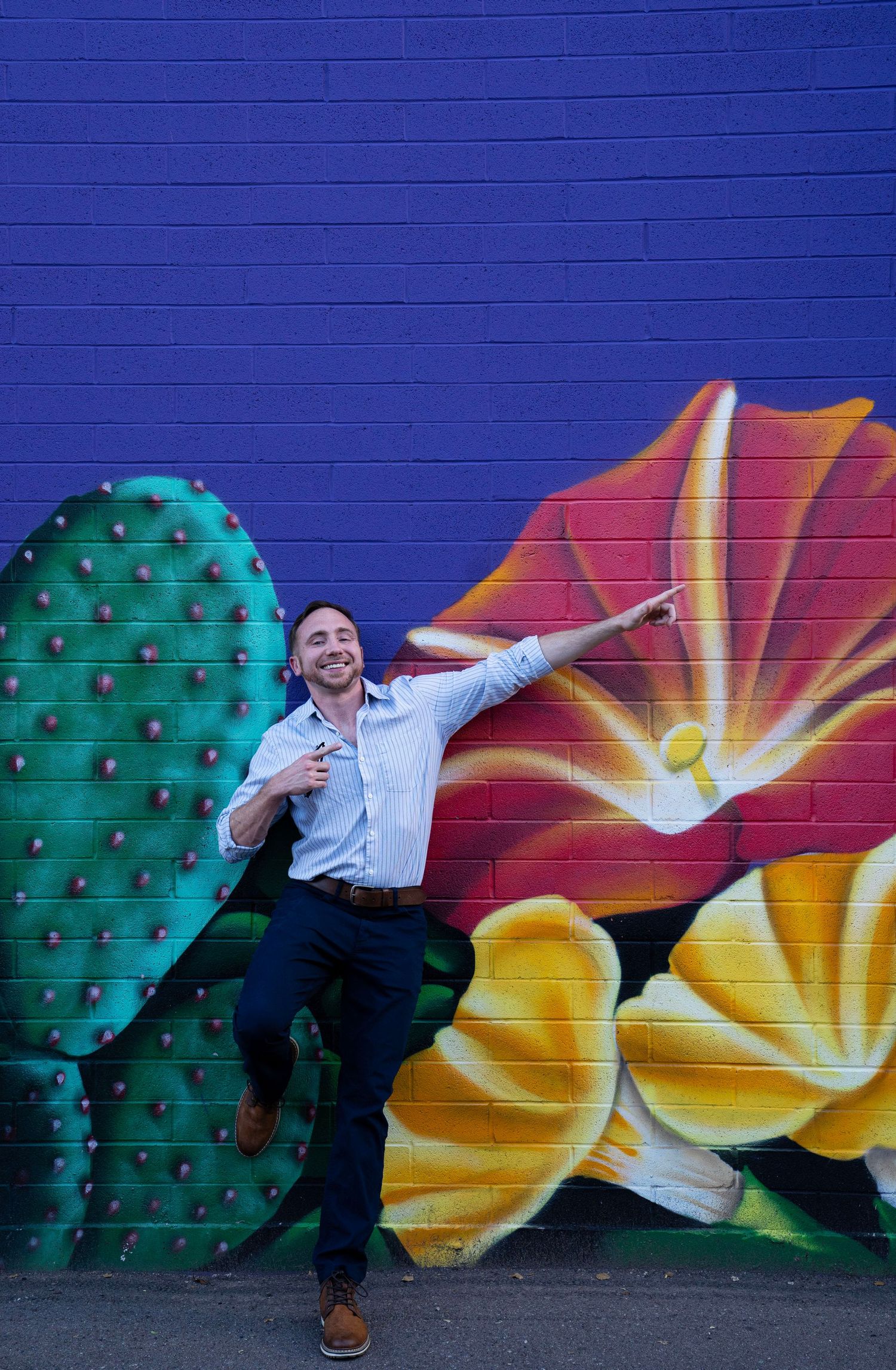 Man posing energetically in front of a vibrant floral and cactus mural on a purple brick wall.