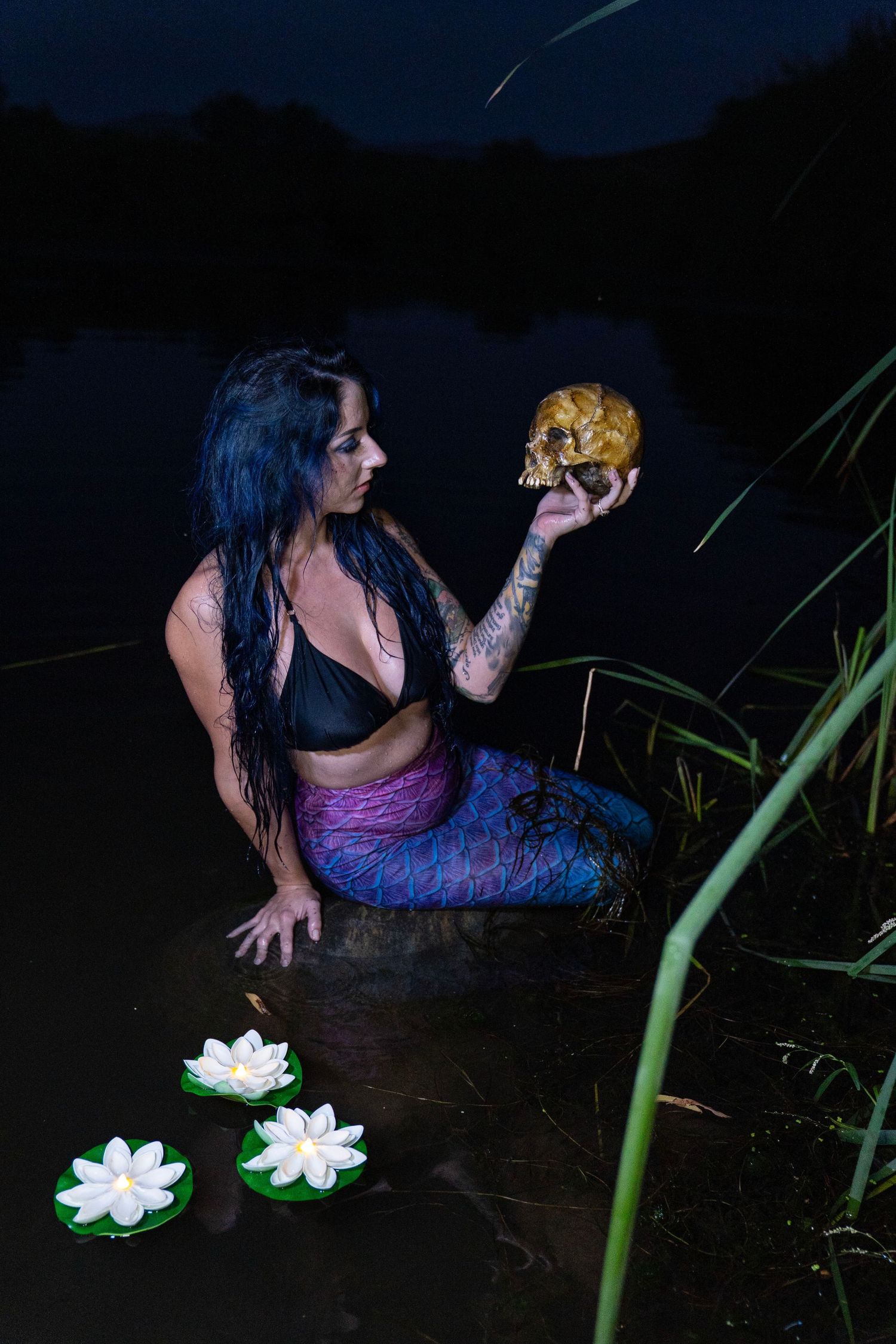 Tattooed woman in mermaid costume holding a skull beside white lotus flowers in dark water at night.