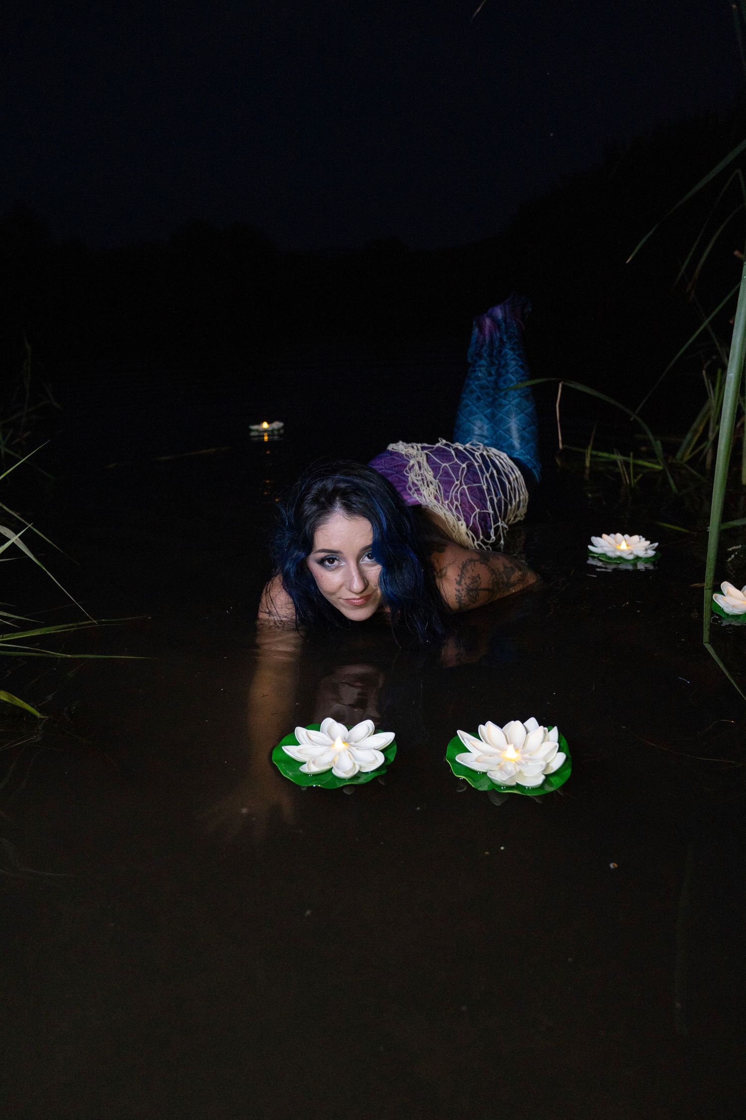 Tattooed woman in mermaid tail lies in dark water beside floating white lotus flowers.