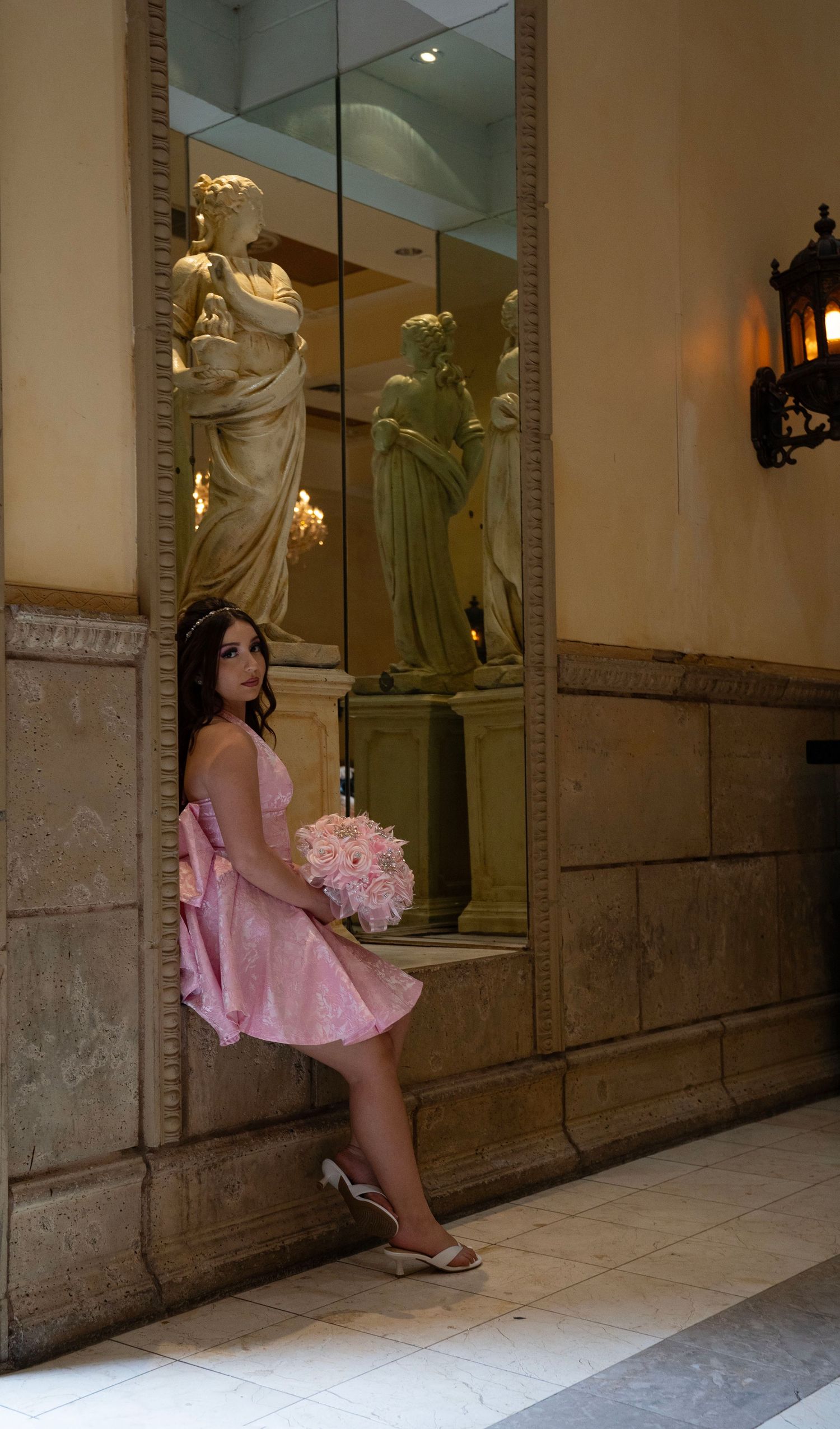 Woman in pink dress holding floral bouquet standing beside ornate mirror with statues.