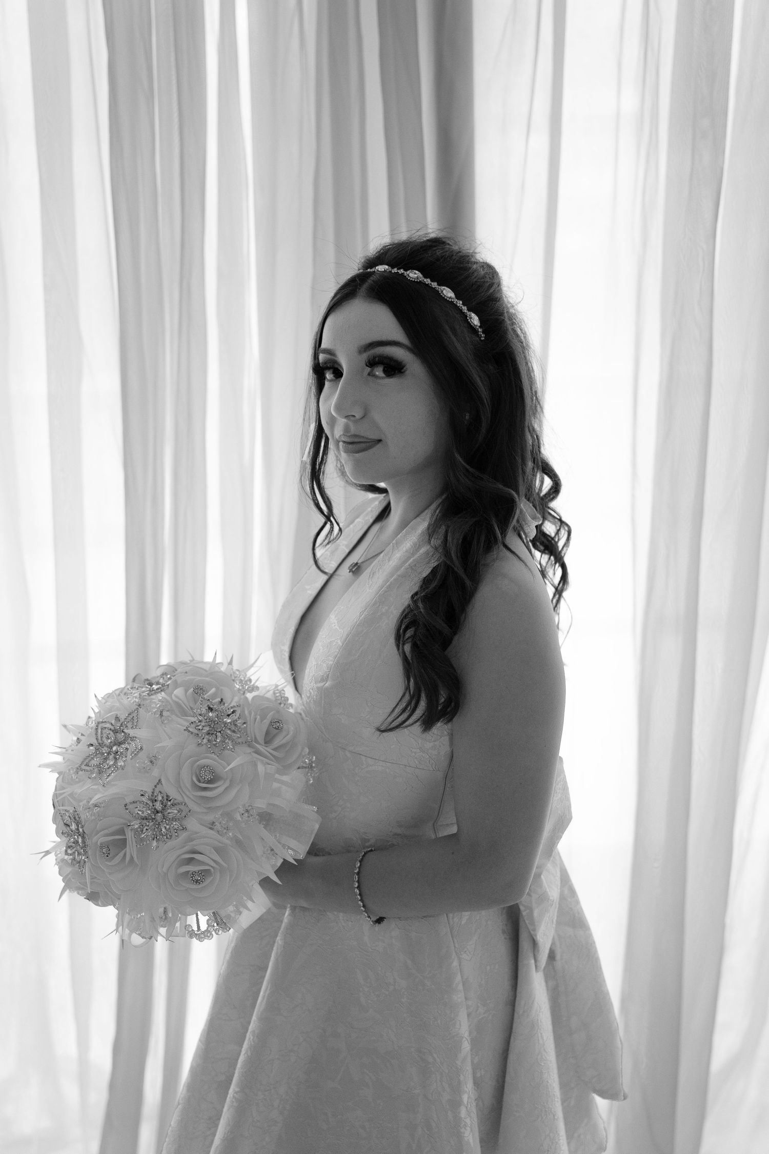 Elegant bride in white gown holding a round floral bouquet with roses, posing in front of sheer white curtains.