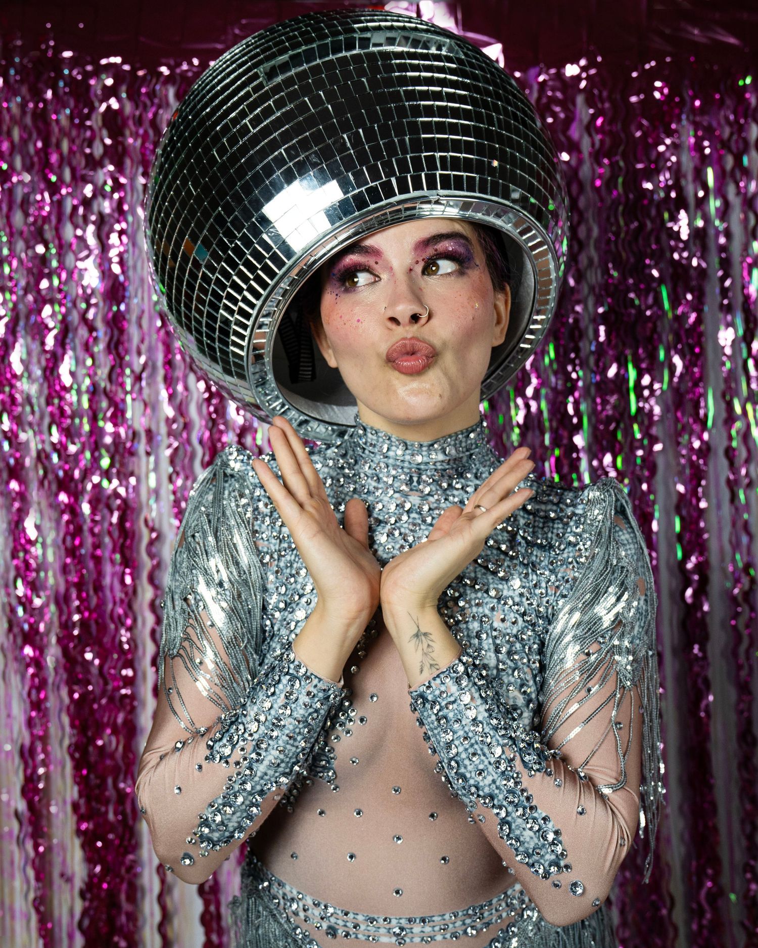 Woman in silver sequin bodysuit wears disco ball helmet, posing against pink tinsel background.