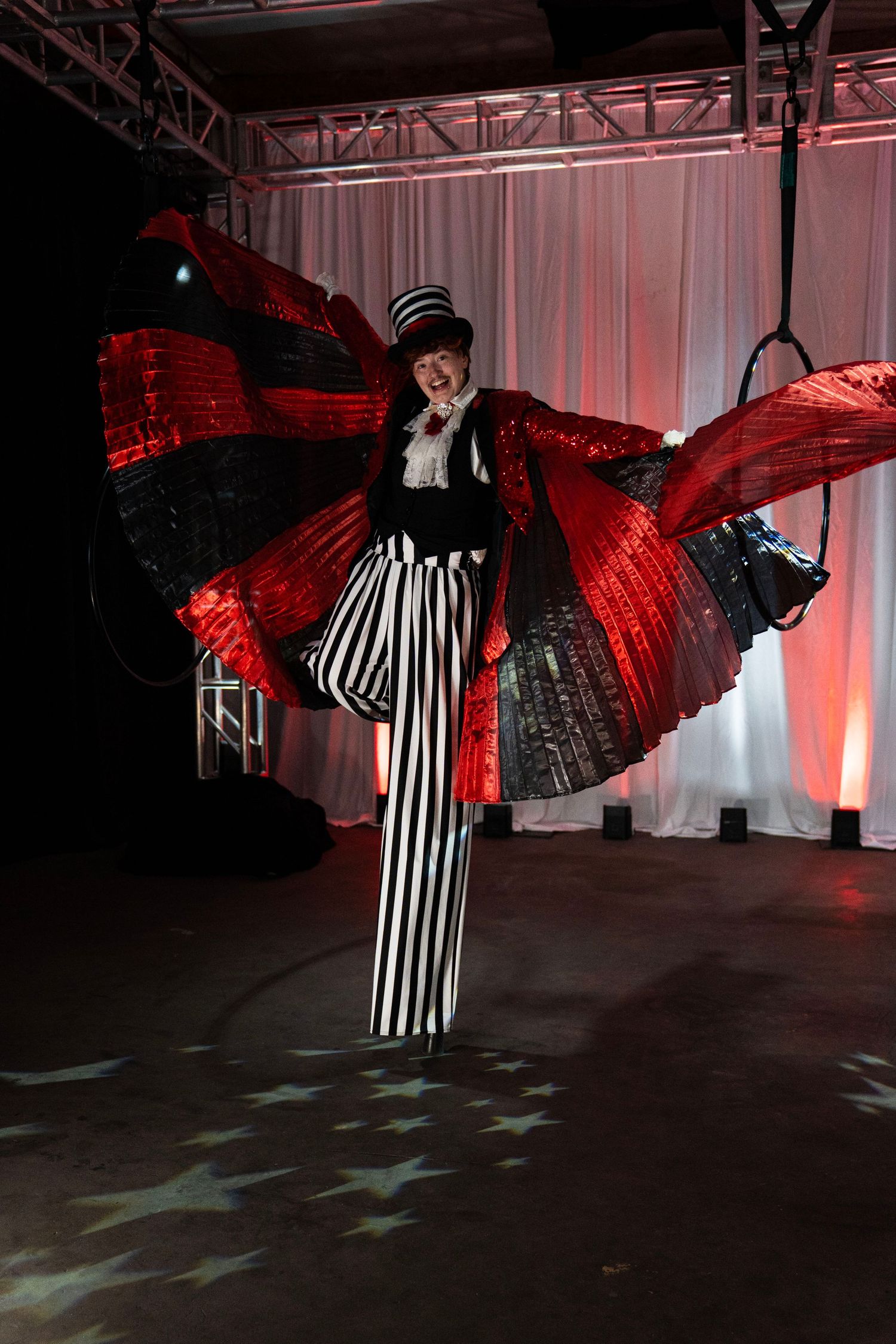 Stilt walker in striped pants and top hat spreads red sequin butterfly wings on stage.