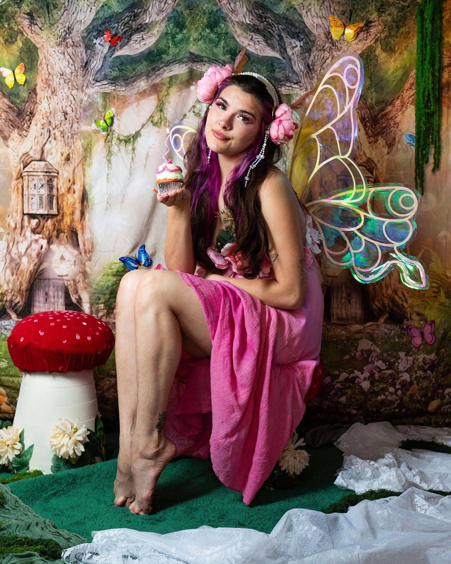 Woman dressed as fairy in pink gown with glowing wings holds cupcake in whimsical woodland photo studio. Fantasy Photoshoot