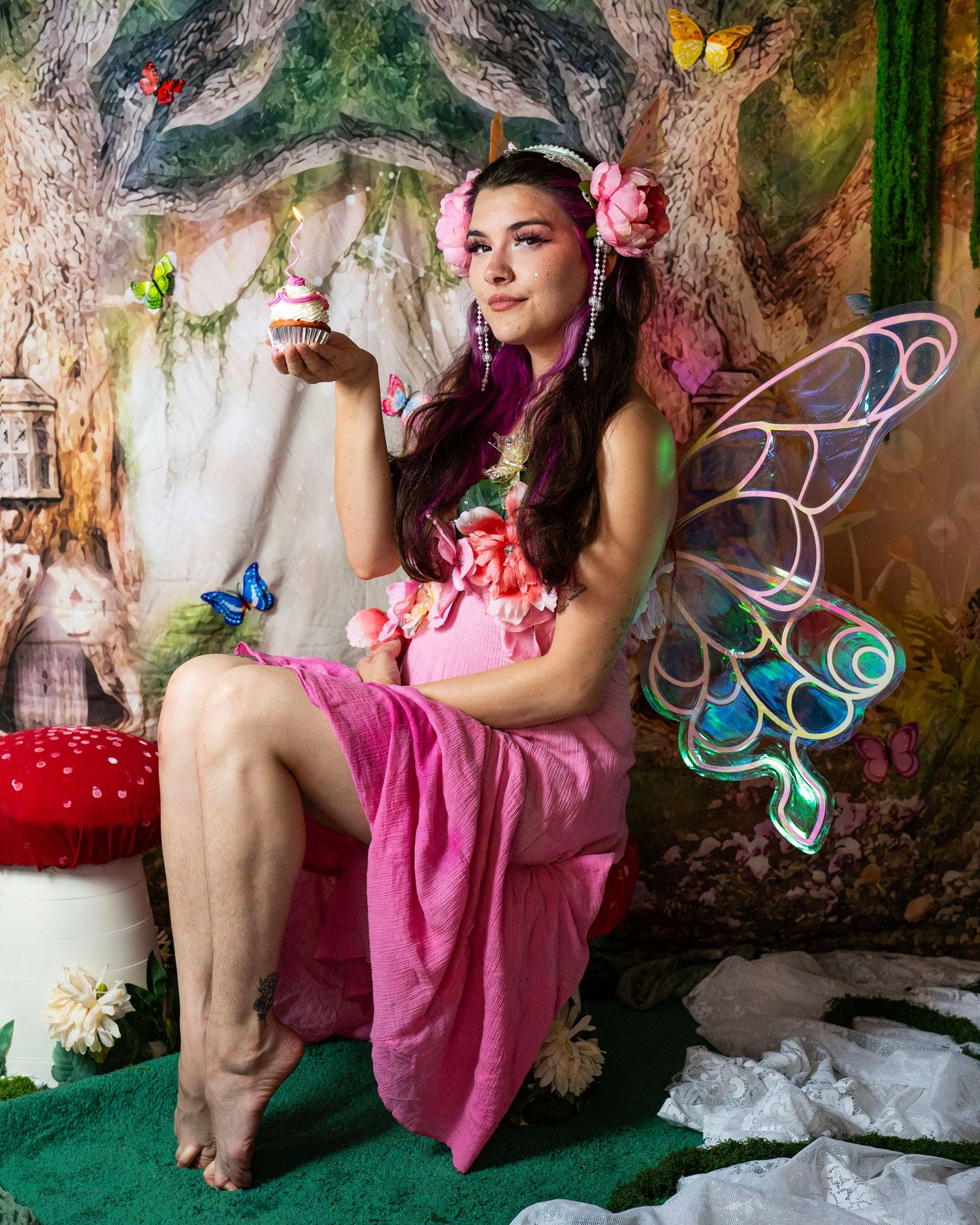 Fairy cosplay model in pink dress with butterfly wings holds cupcake in whimsical enchanted forest setting. Fantasy Photoshoot