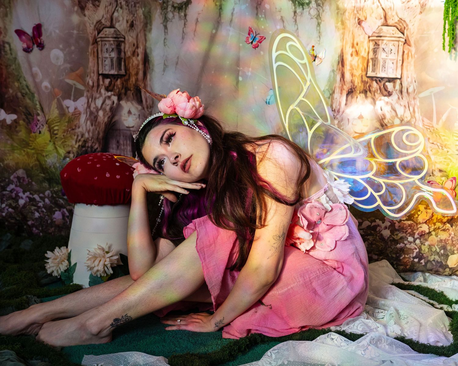 Young woman in pink fairy costume with wings resting in a magical forest photography set. Fantasy Photoshoot