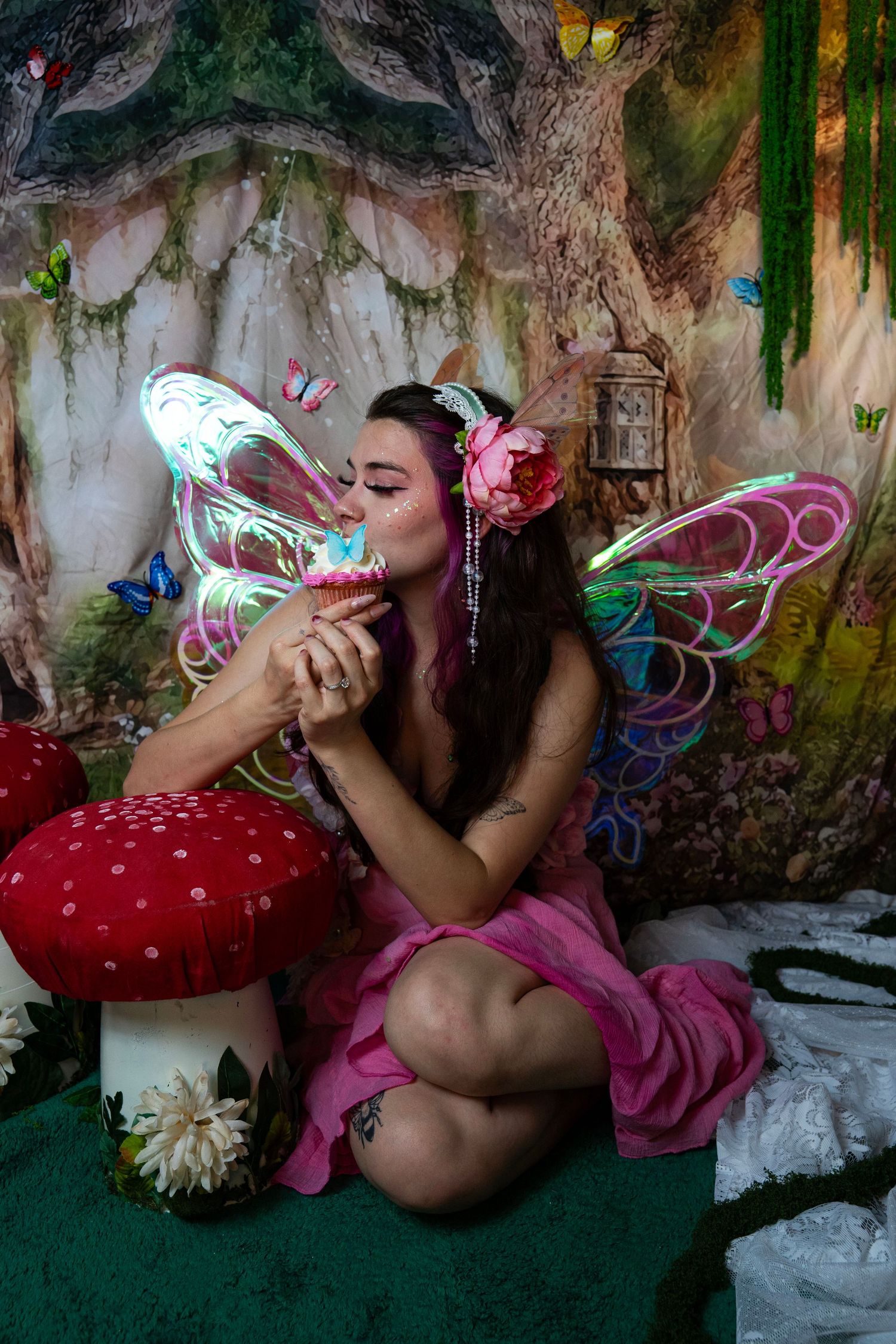 Woman in pink fairy costume with iridescent wings sipping from a small cup in enchanted forest backdrop. Fantasy Photoshoot