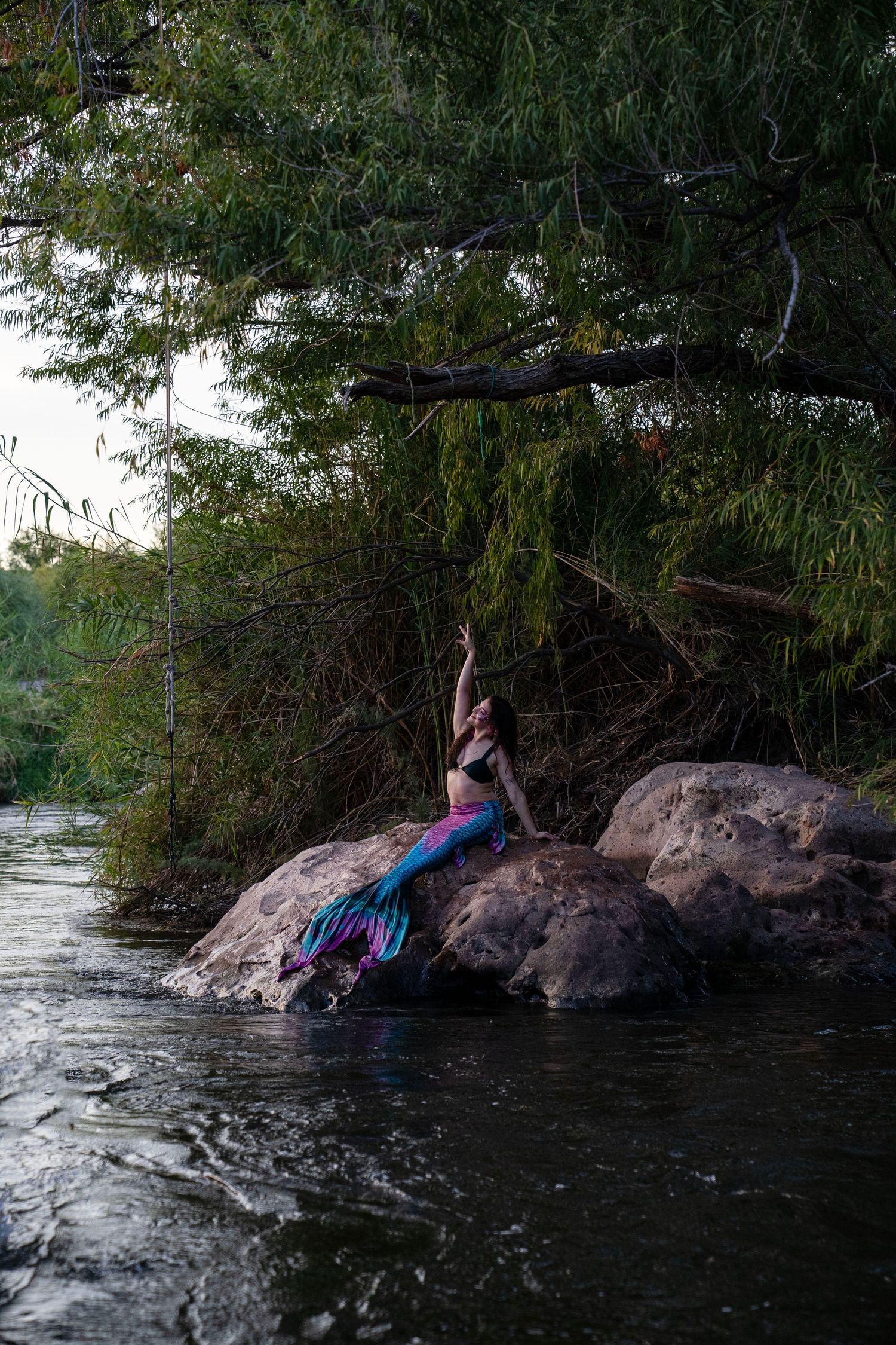 A woman in a colorful mermaid tail arches gracefully on rocks beside a river surrounded by lush green trees. Fantasy Photoshoot