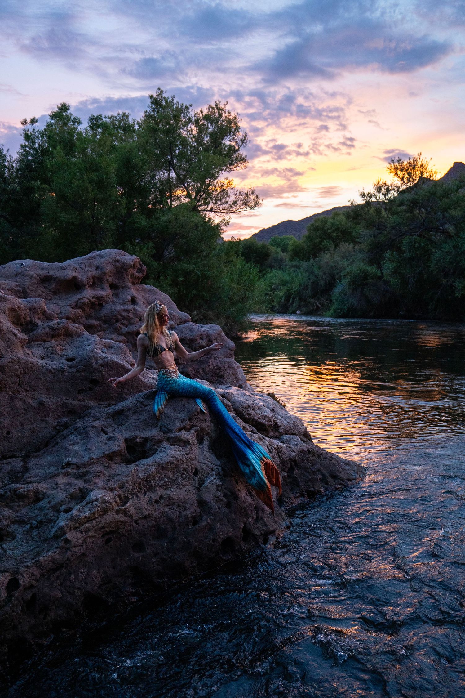 Woman in blue mermaid tail costume sitting on rocky riverbank at dusk. Mermaid photoshoot, fantasy photoshoot. Arizona photographer.
