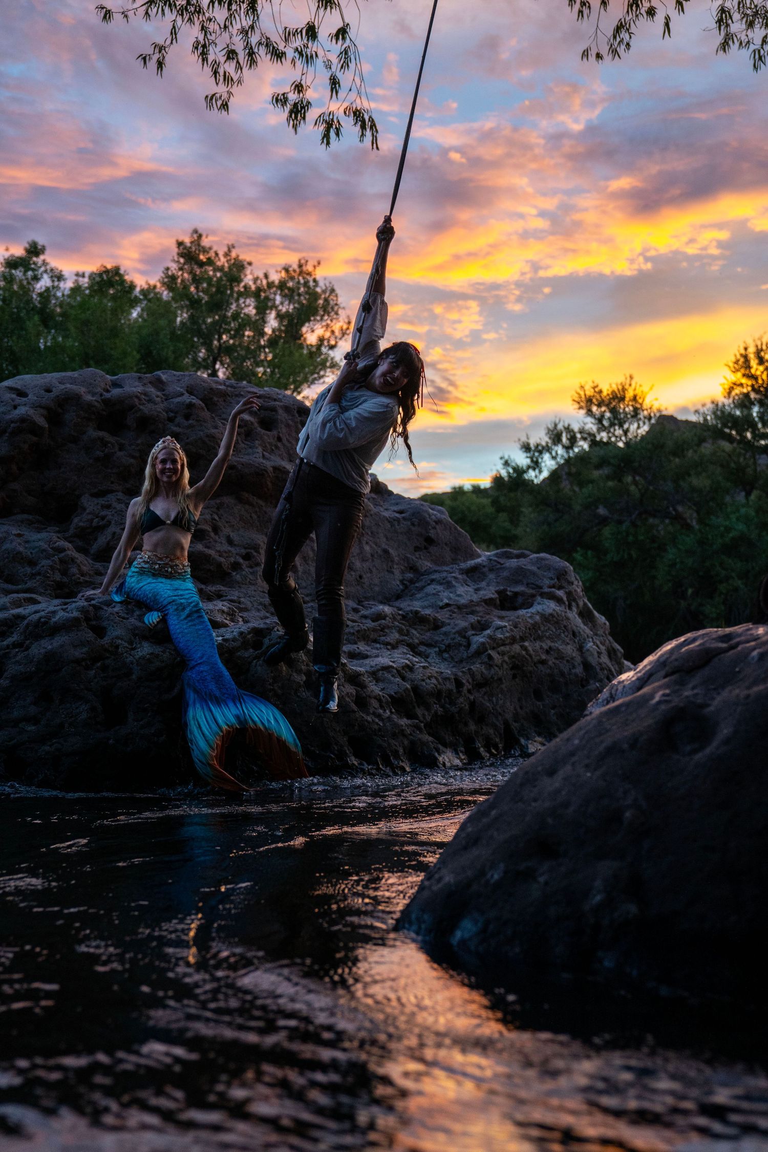 Pirate and mermaid pose on rocks by river at colorful sunset. Mermaid photoshoot, fantasy photoshoot. Arizona photographer.