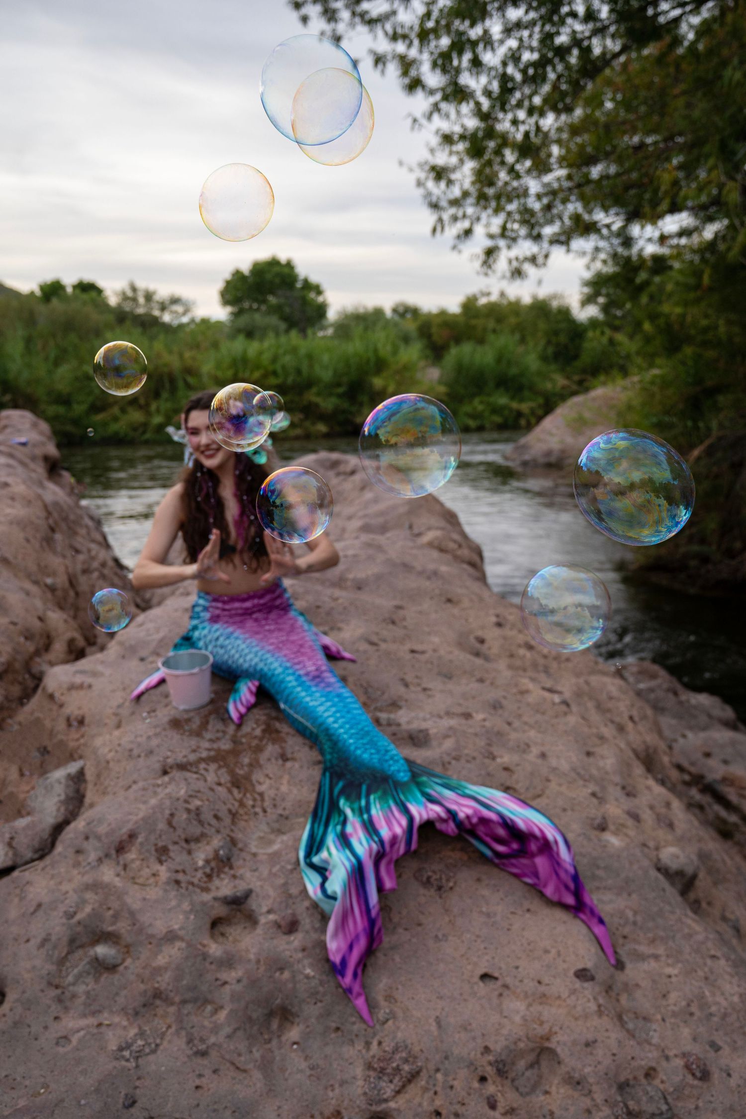A woman in a vibrant mermaid tail costume sits on rocks surrounded by large floating soap bubbles. Fantasy Photoshoot