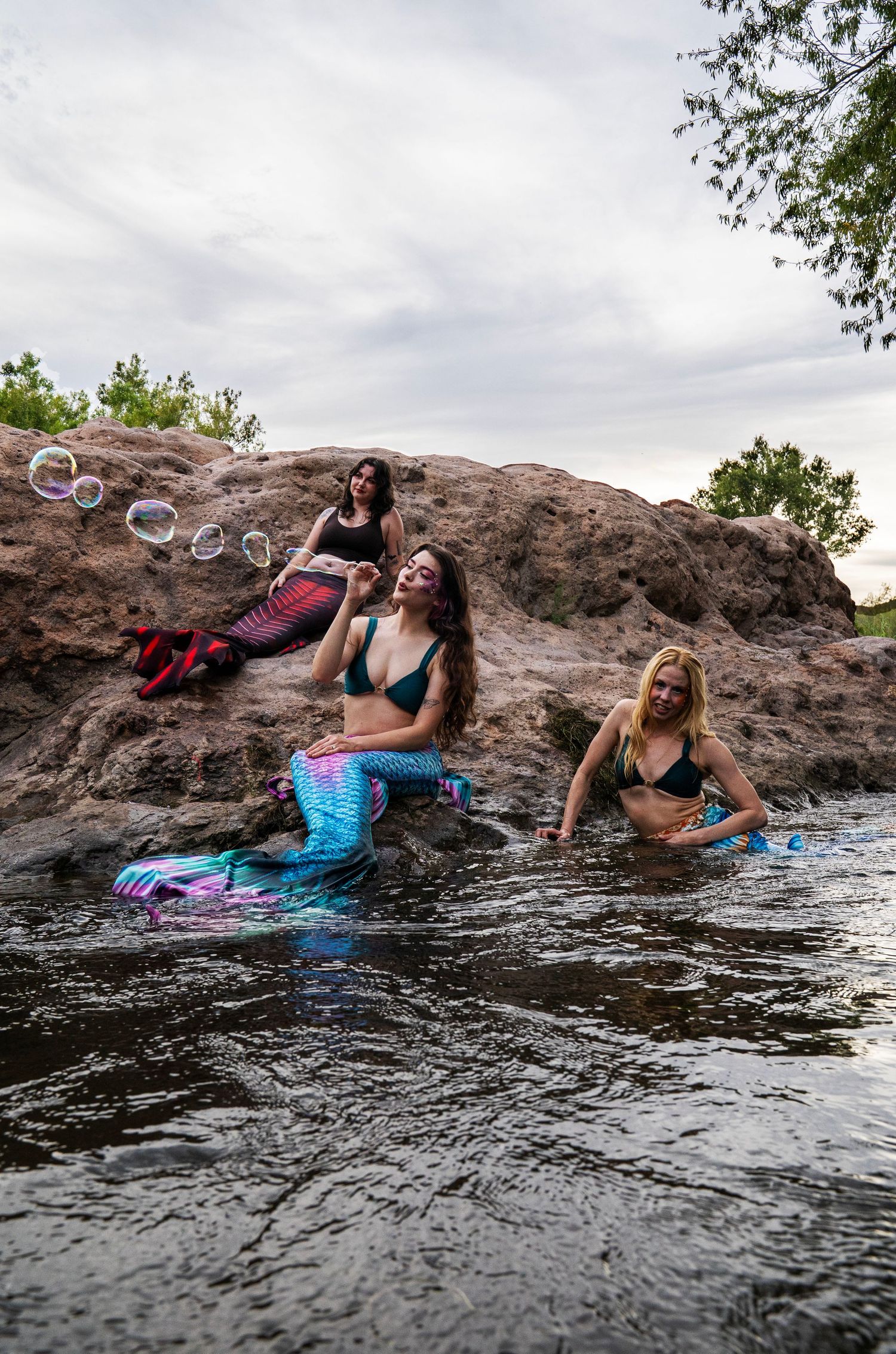 Three women in mermaid costumes play with bubbles on rocky riverbank surrounded by lush greenery. Fantasy Photoshoot
