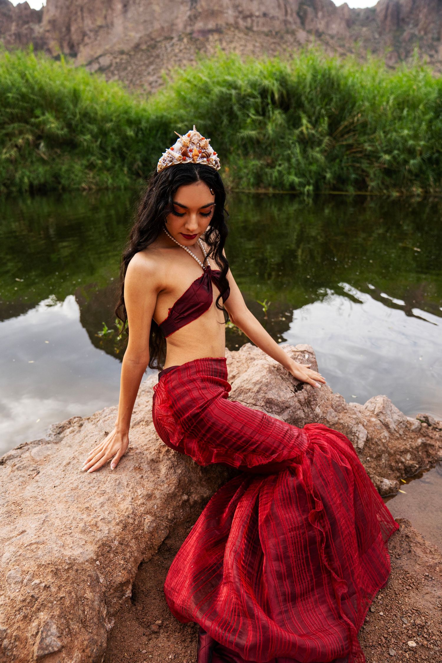 A woman in a red mermaid tail and shell crown sits gracefully on rocks beside a calm green river.