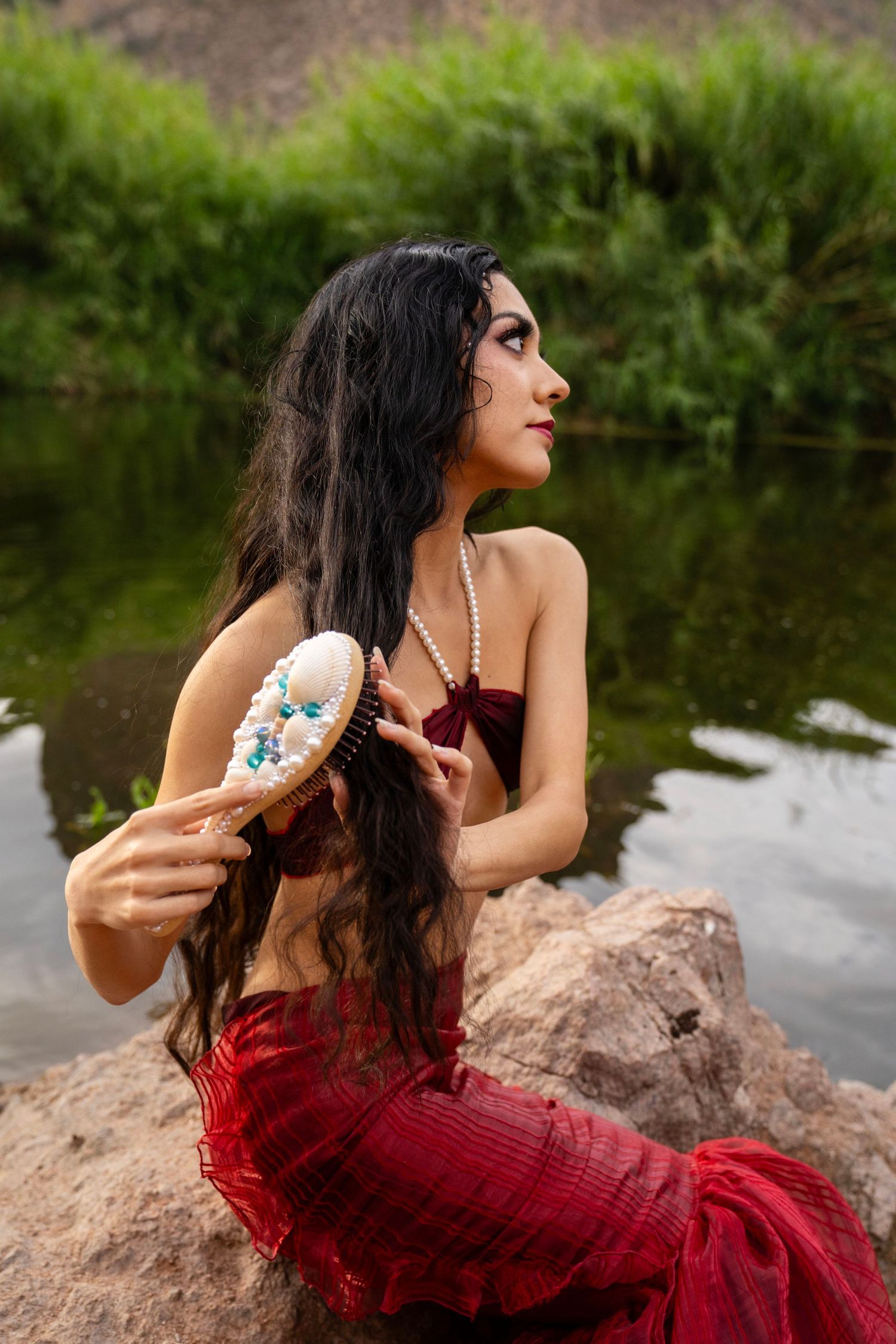 Woman dressed as a mermaid in red tail and pearl necklace brushing hair on rocks by a river.