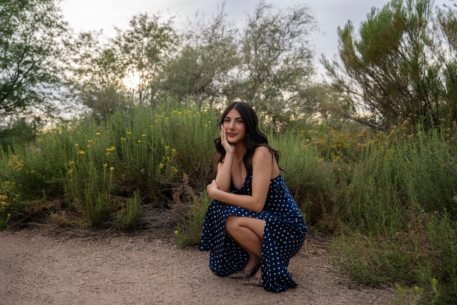 Young woman in navy polka dot dress crouching on desert trail surrounded by lush green shrubs and wildflowers at sunset.