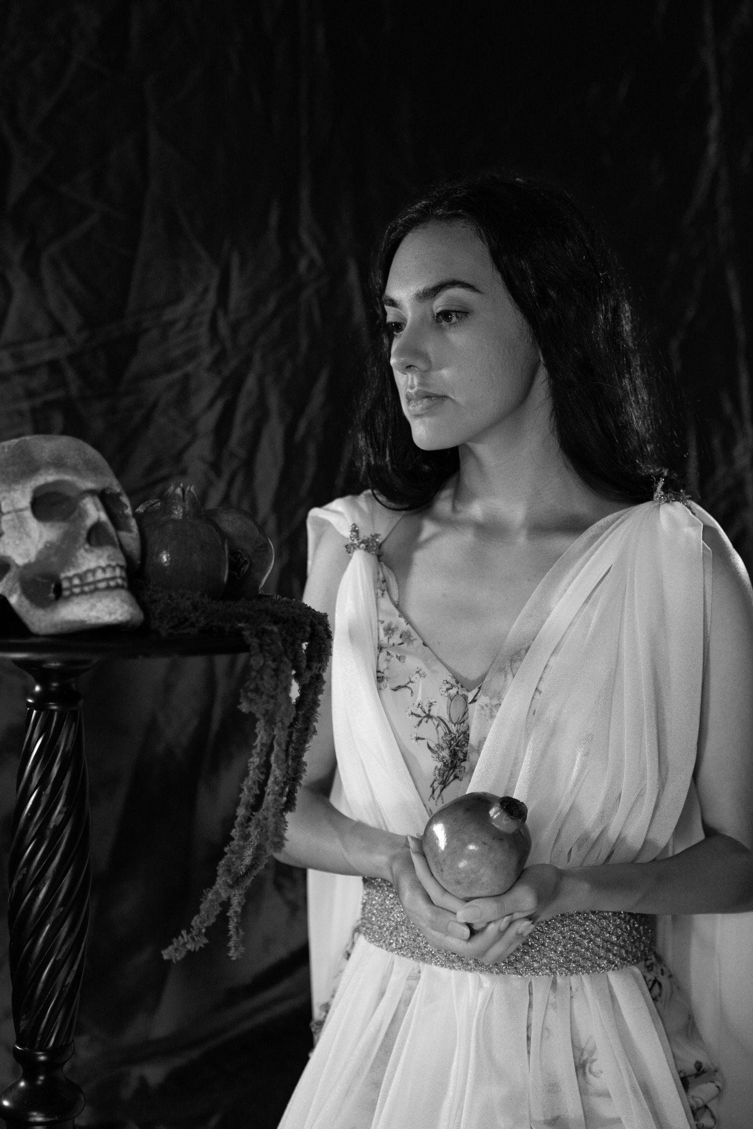 Black and white portrait of a woman in a flowing floral gown holding a pomegranate beside a human skull on an ornate table.