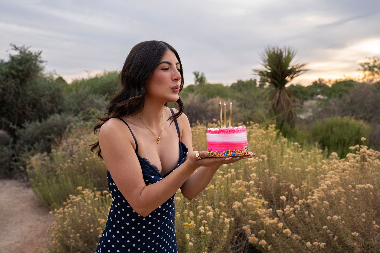 Young brunette woman in a polka dot dress blowing out candles on a pink birthday cake outdoors.