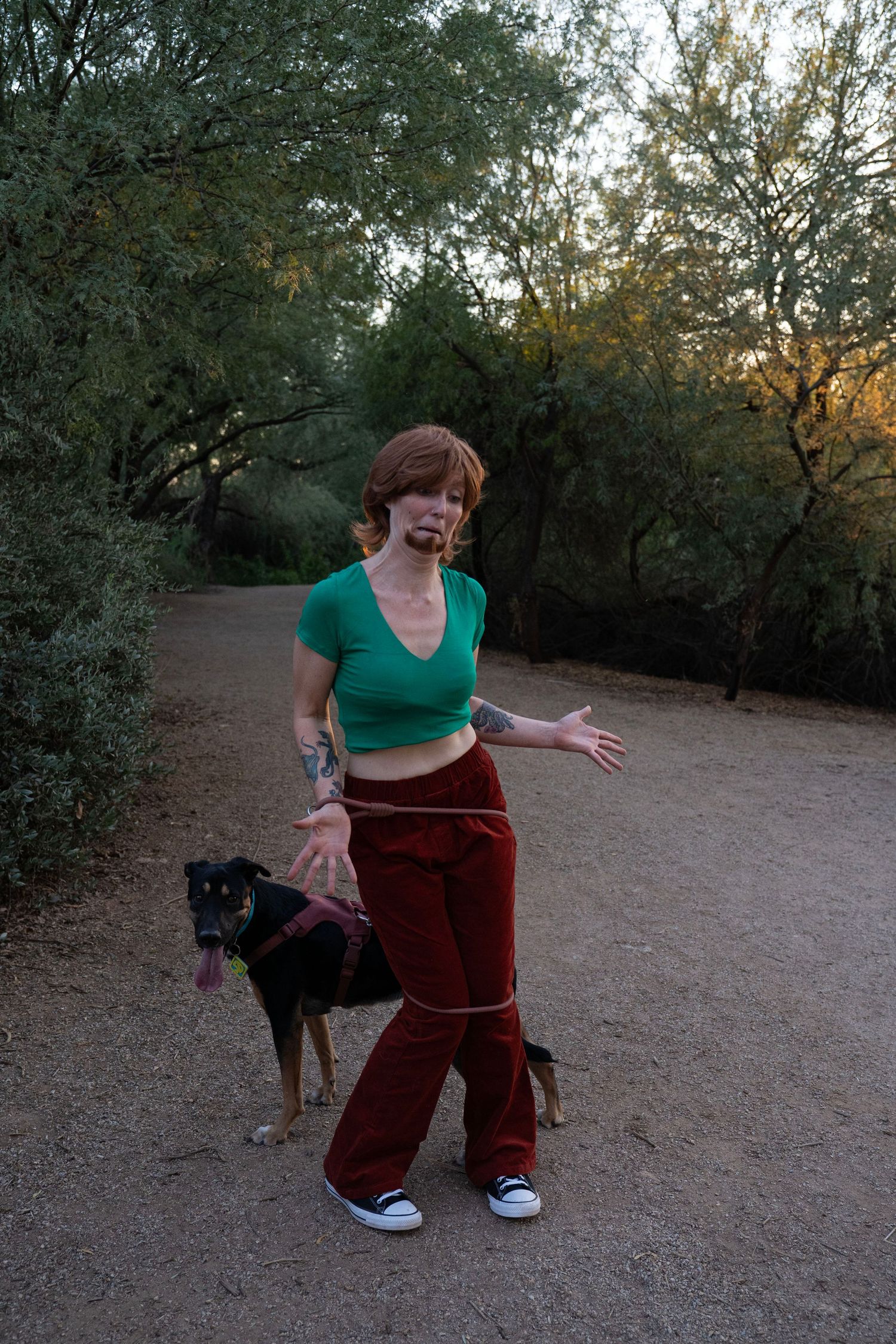 A red-haired woman in a green crop top and maroon pants stands on a dirt trail with a black and tan dog on a leash.