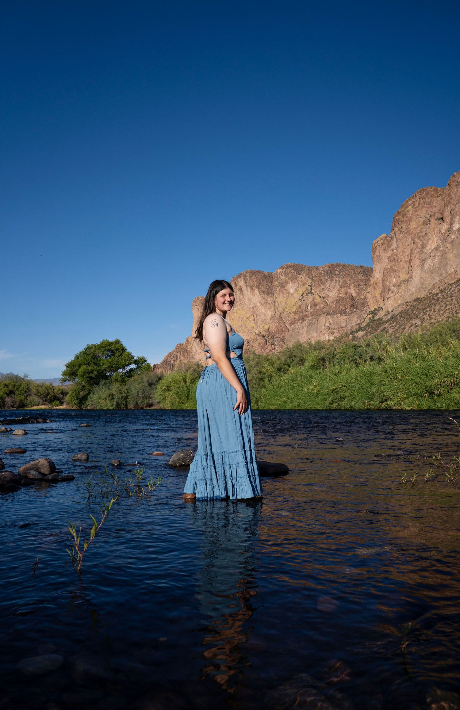 Woman in blue maxi dress standing in a shallow river with rocky cliffs and blue sky backdrop.
