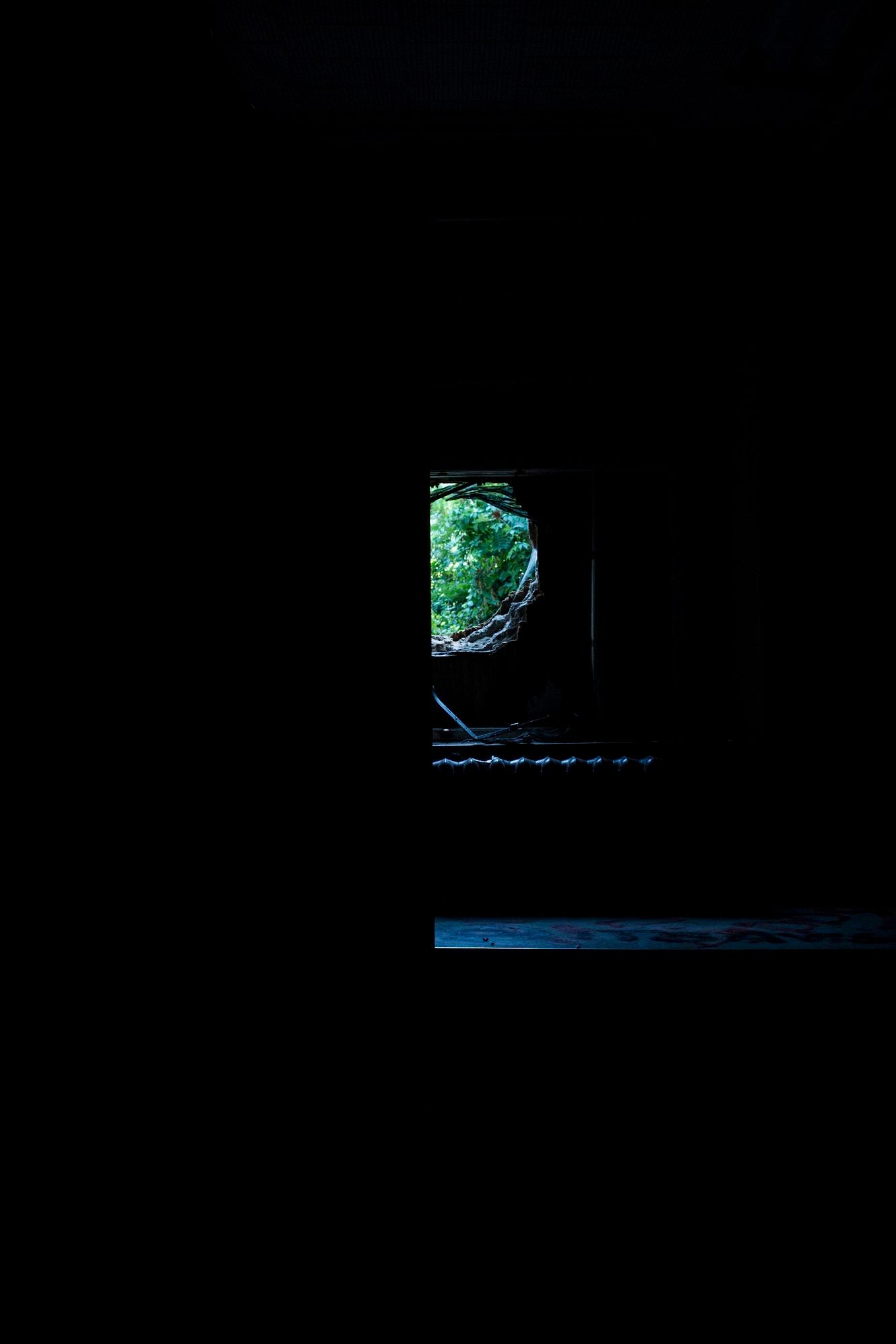 Dark interior of an old building with a small lit window revealing lush green foliage and sunlight outside.