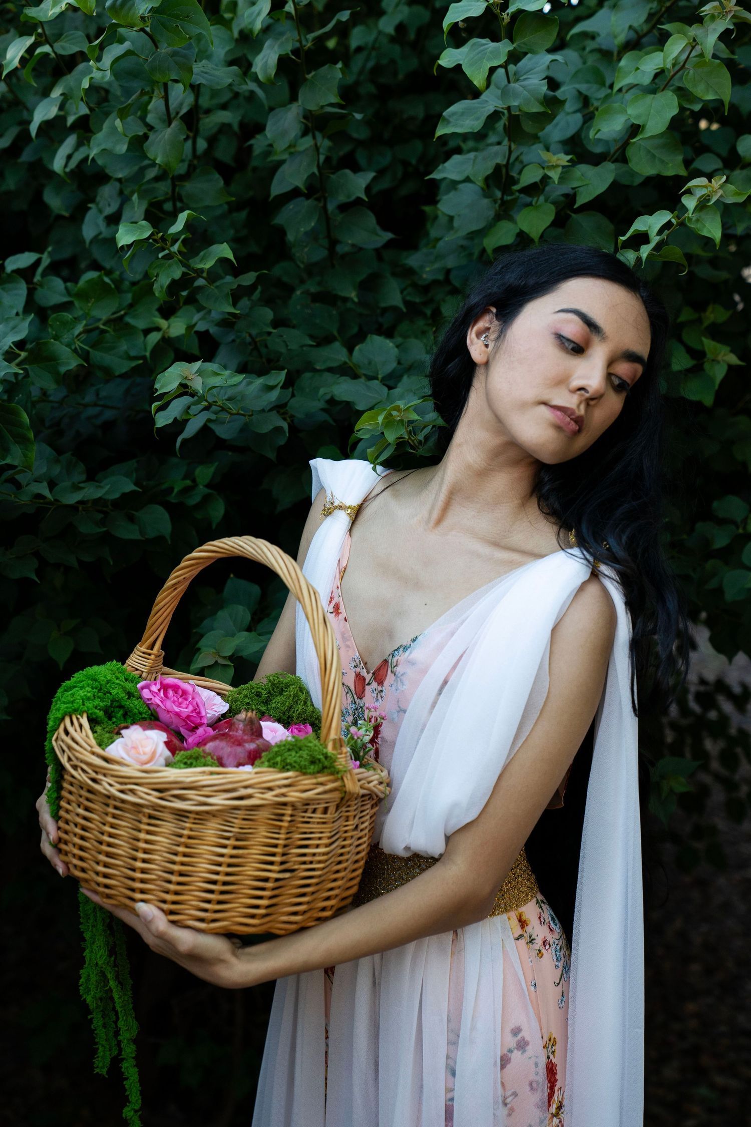 Woman in a flowing pink floral dress with closed eyes holds a wicker basket of pink roses and moss before lush green foliage.