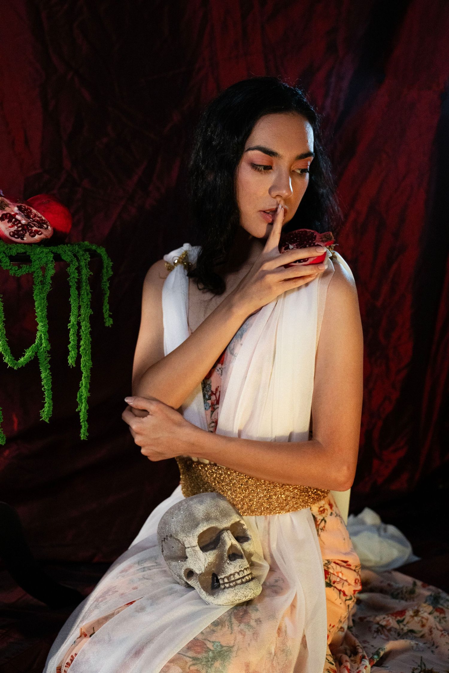 Woman in a white draped gown with gold belt poses with finger to lips beside a skull and pomegranate against deep red fabric.