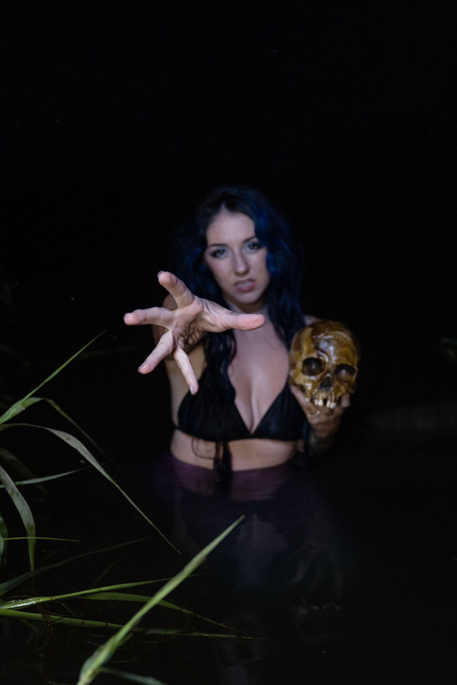 Dark-haired woman in a black bikini top stands waist-deep in night water, reaching toward the camera while holding a golden skull.