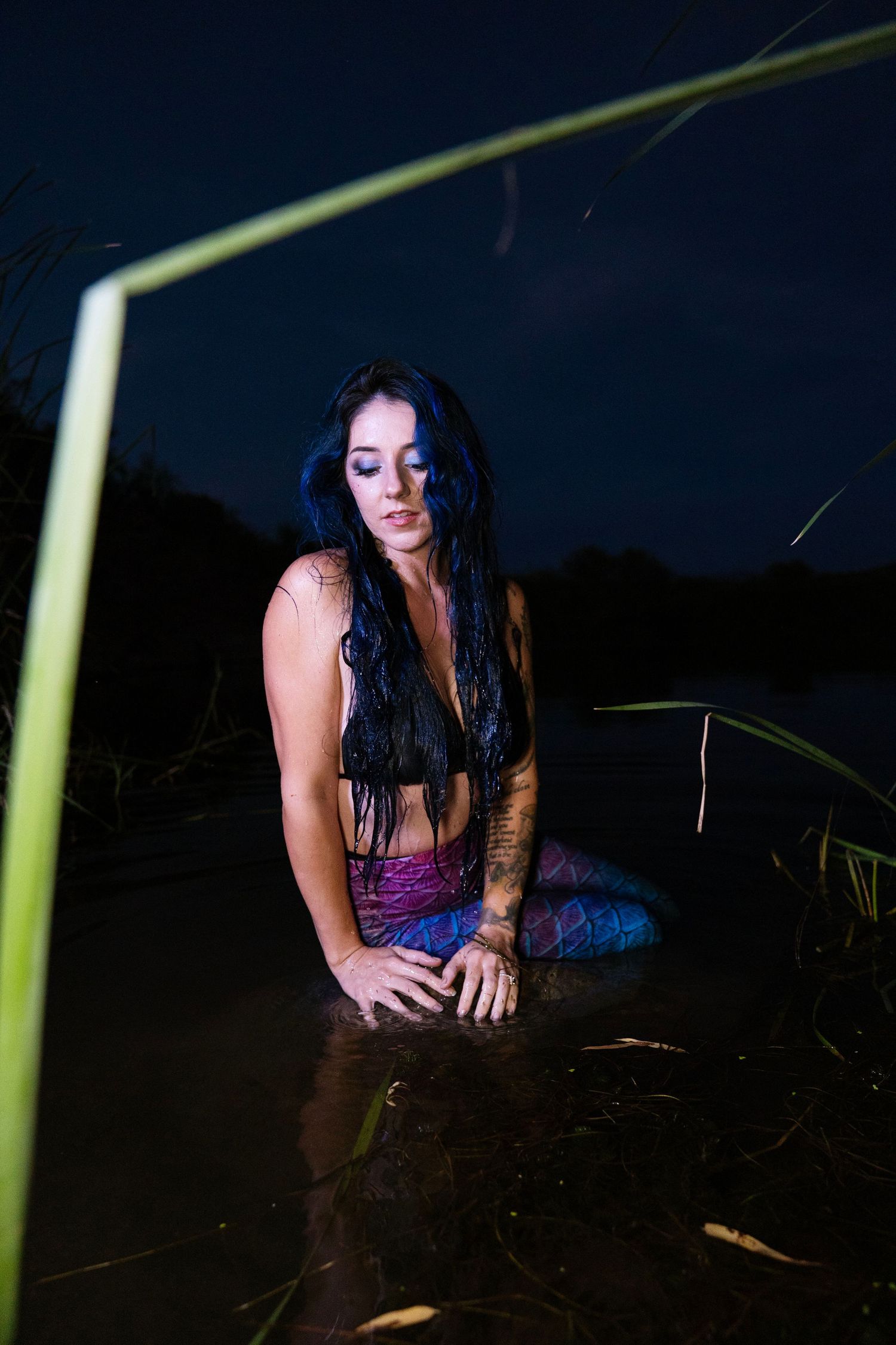 Woman with long dark wet hair kneeling in shallow water at night near tall grass.