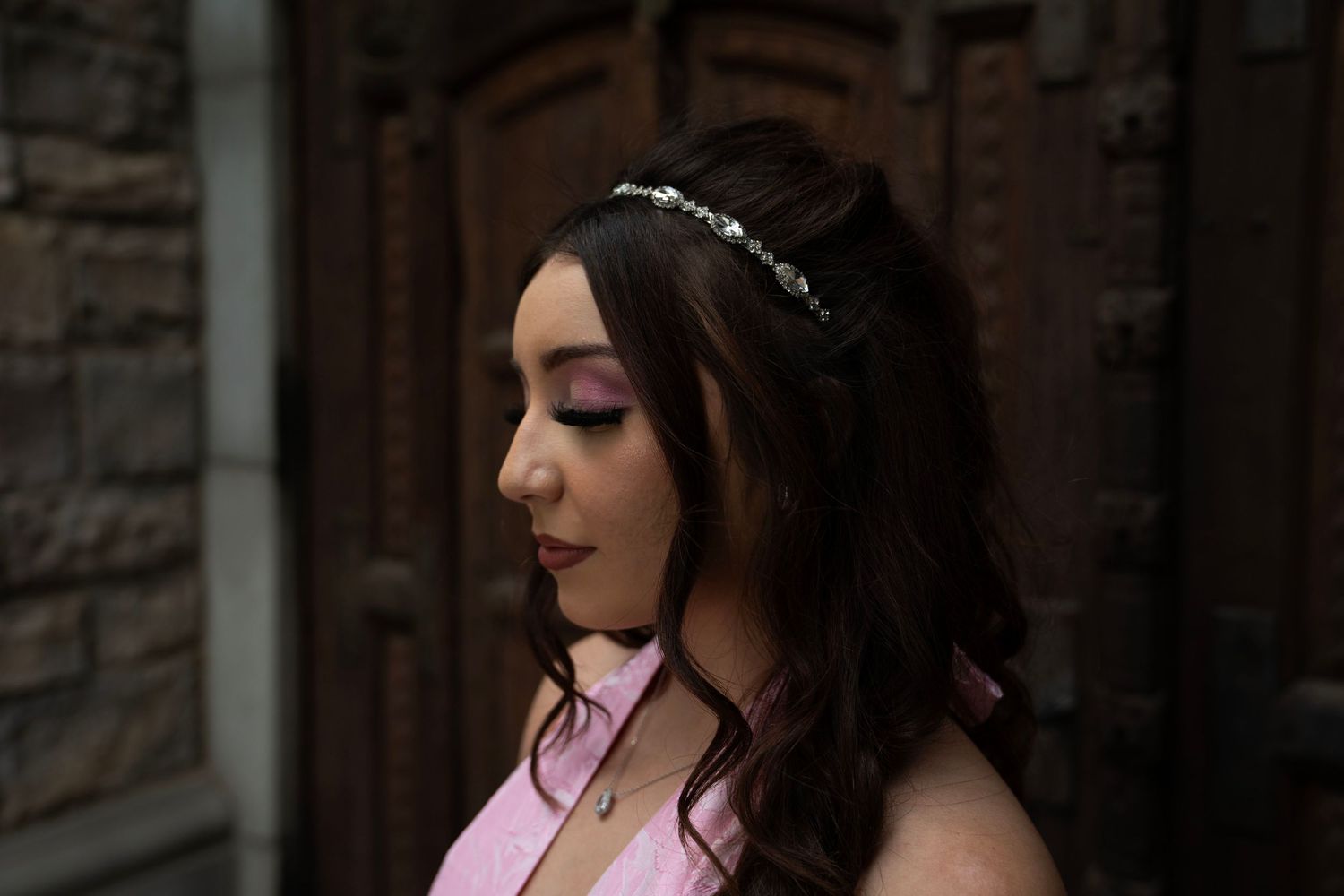 Young woman in pink dress with floral headband and purple eyeshadow posing elegantly outdoors.