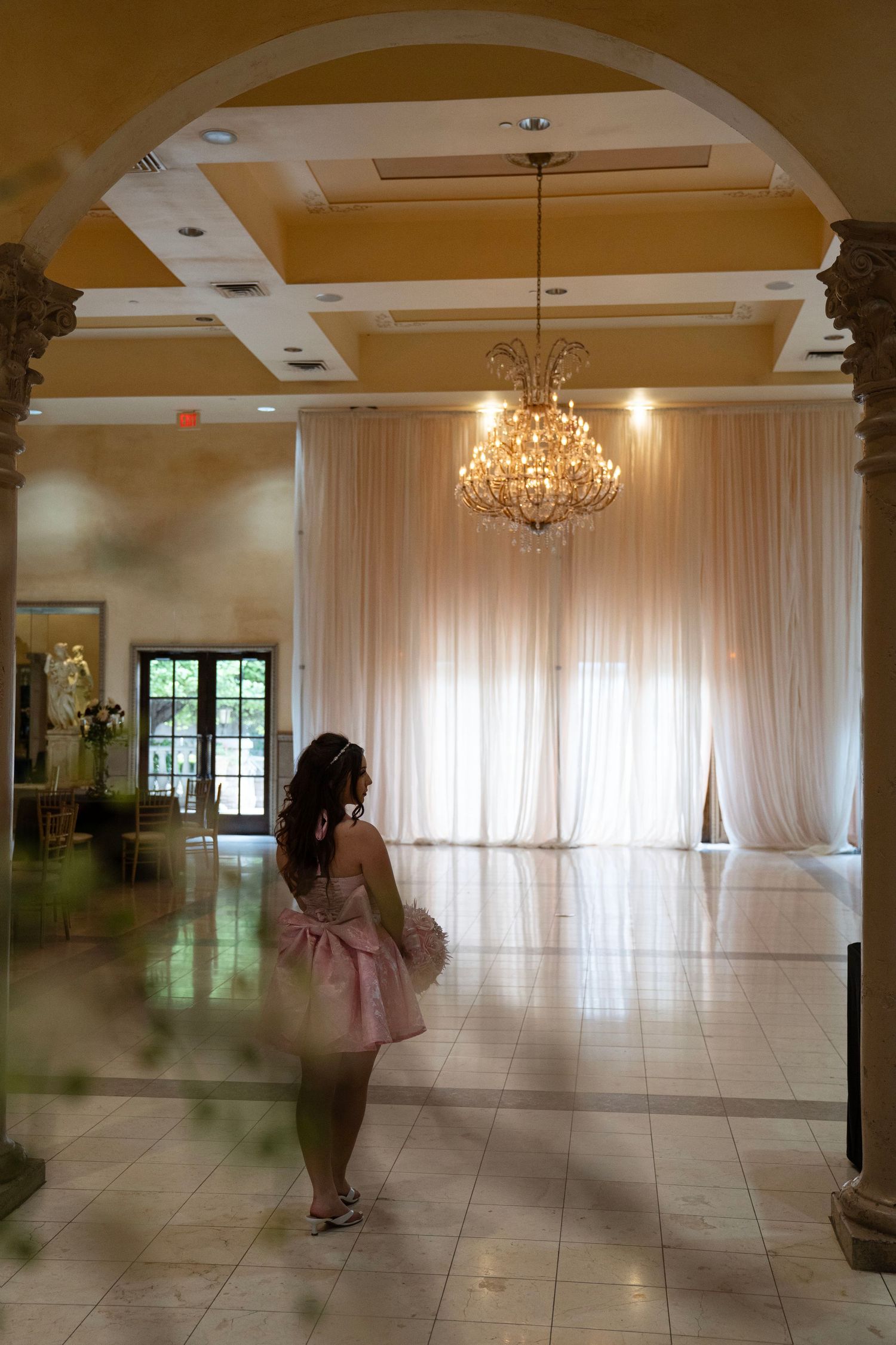 Woman in pink strapless dress standing in elegant venue hallway beneath a crystal chandelier with white draping.