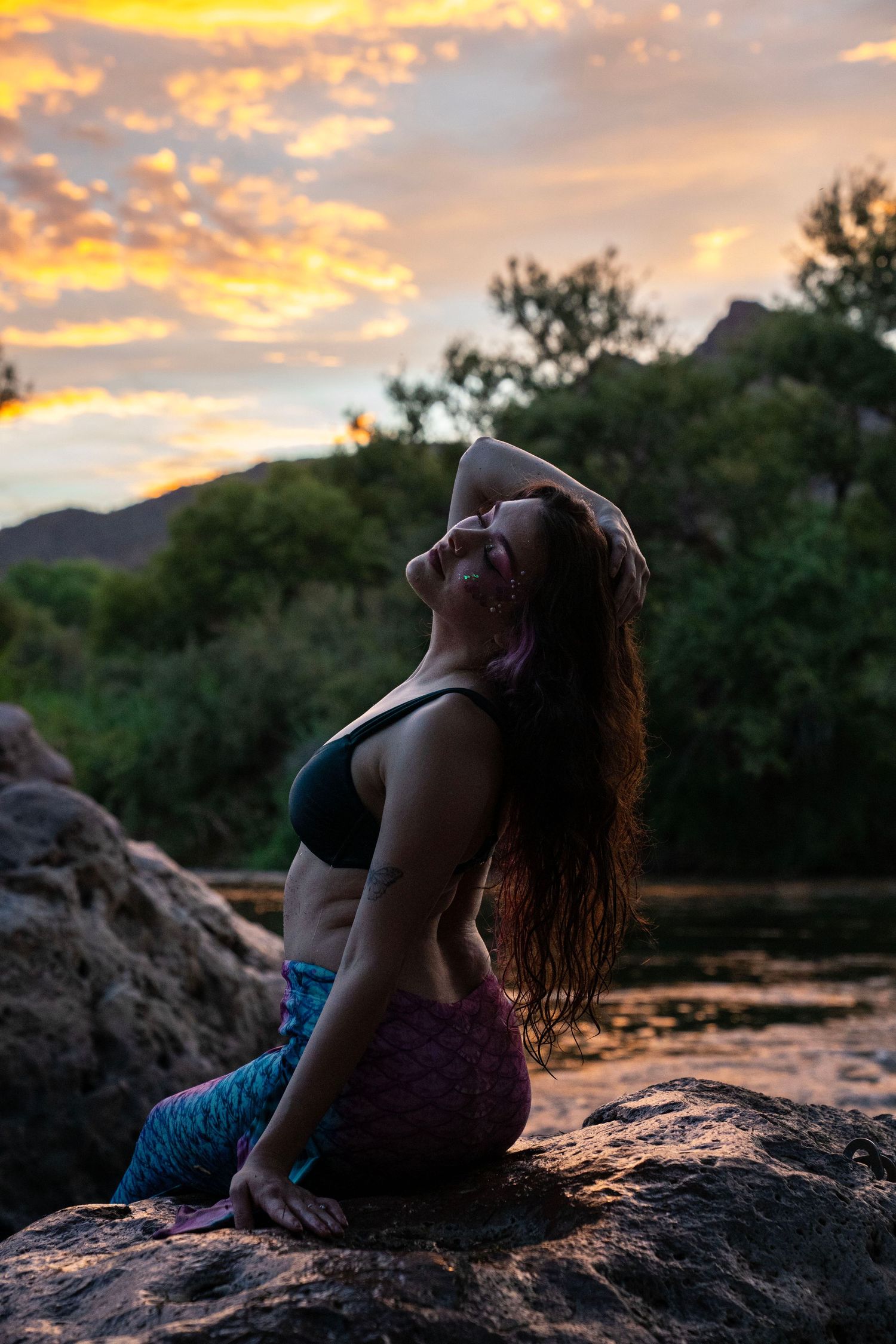 A smiling woman in a mermaid costume arches back on riverside rocks beneath a dramatic sunset sky. Mermaid photoshoot, fantasy photoshoot.