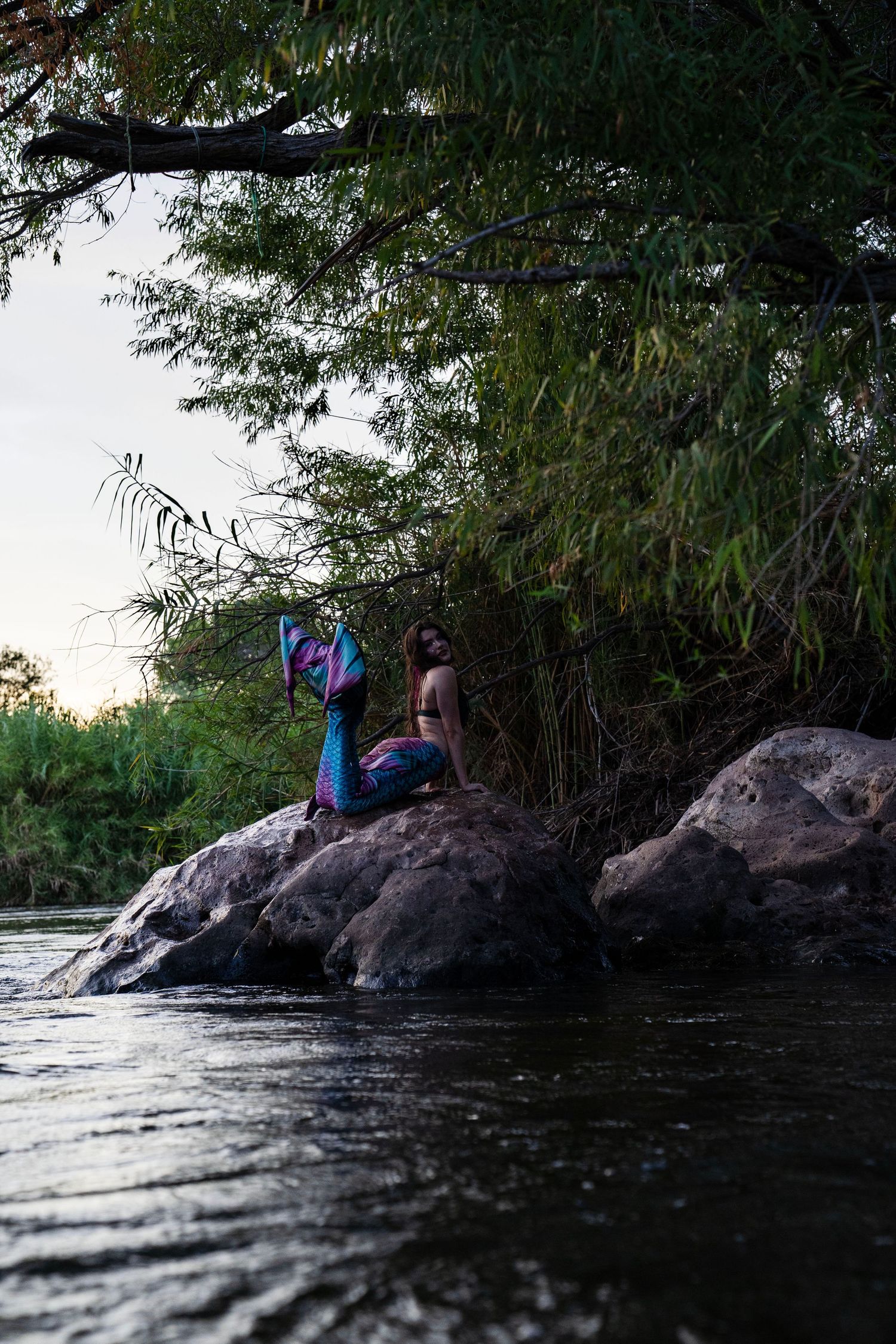 A woman wearing a vibrant mermaid tail sits on large rocks by a river, surrounded by lush green trees at dusk. Fantasy Photoshoot