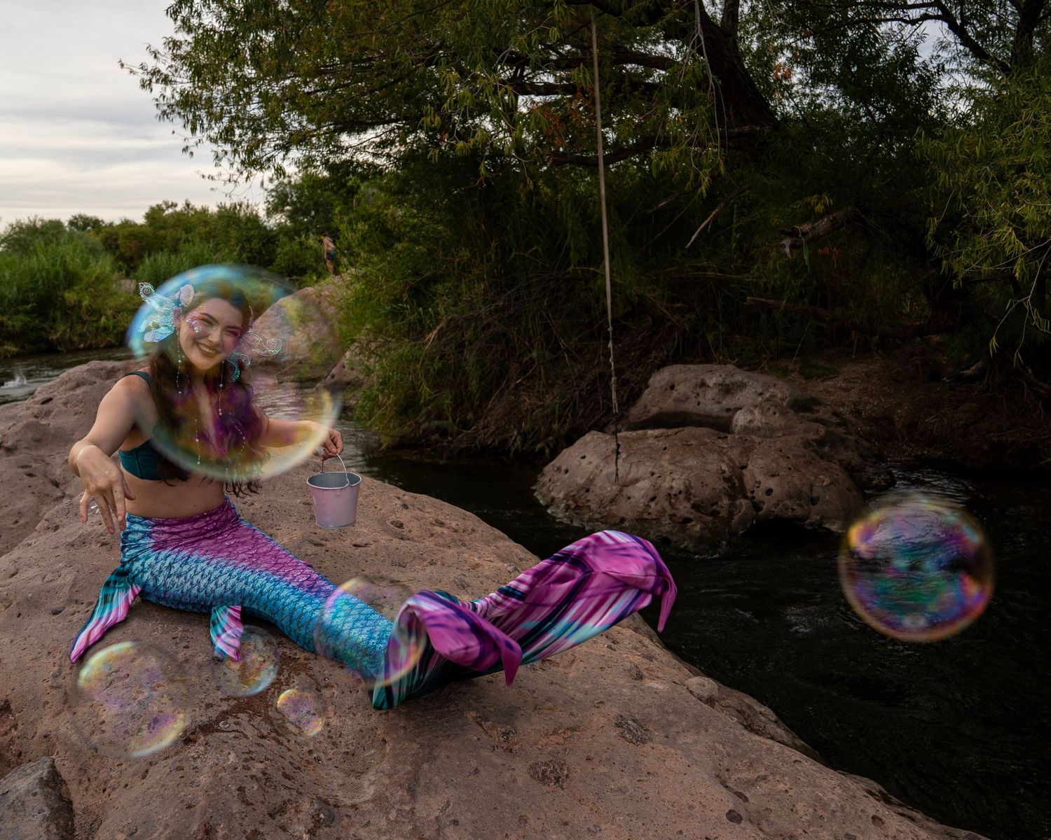 Woman in mermaid costume with blue wig playing with giant soap bubbles while sitting on river rocks. Fantasy Photoshoot