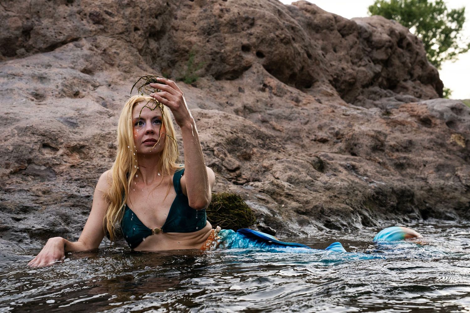 A blonde woman dressed as a mermaid with a blue tail sits in shallow river water near rocky shores. Fantasy Photoshoot