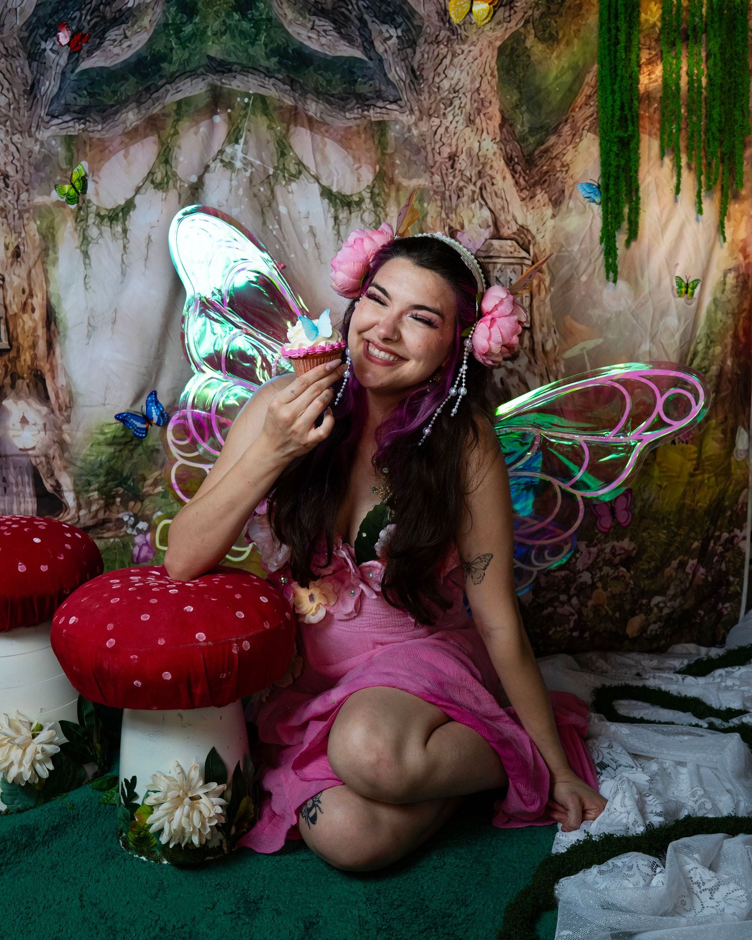Smiling woman in pink fairy costume with iridescent wings posing near decorative mushrooms in enchanted forest. Fantasy Photoshoot