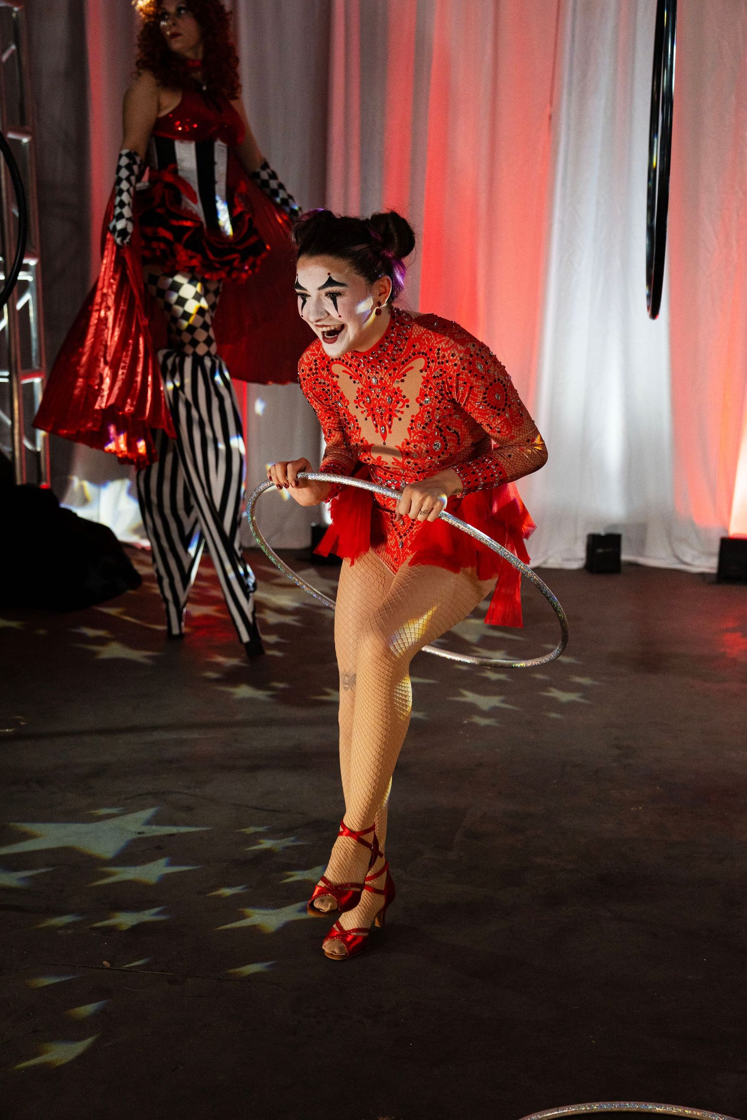 Circus performer with clown makeup spins hula hoop in red sequin costume on star-lit stage.