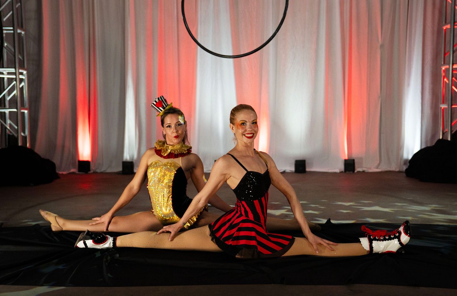 Two circus performers in colorful costumes doing splits pose in front of aerial hoop.