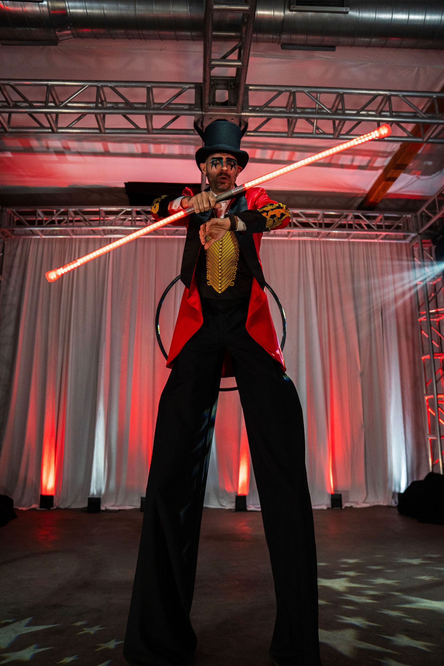 Circus ringmaster on stilts holds glowing red batons in a powerful stance on a dramatically lit stage.