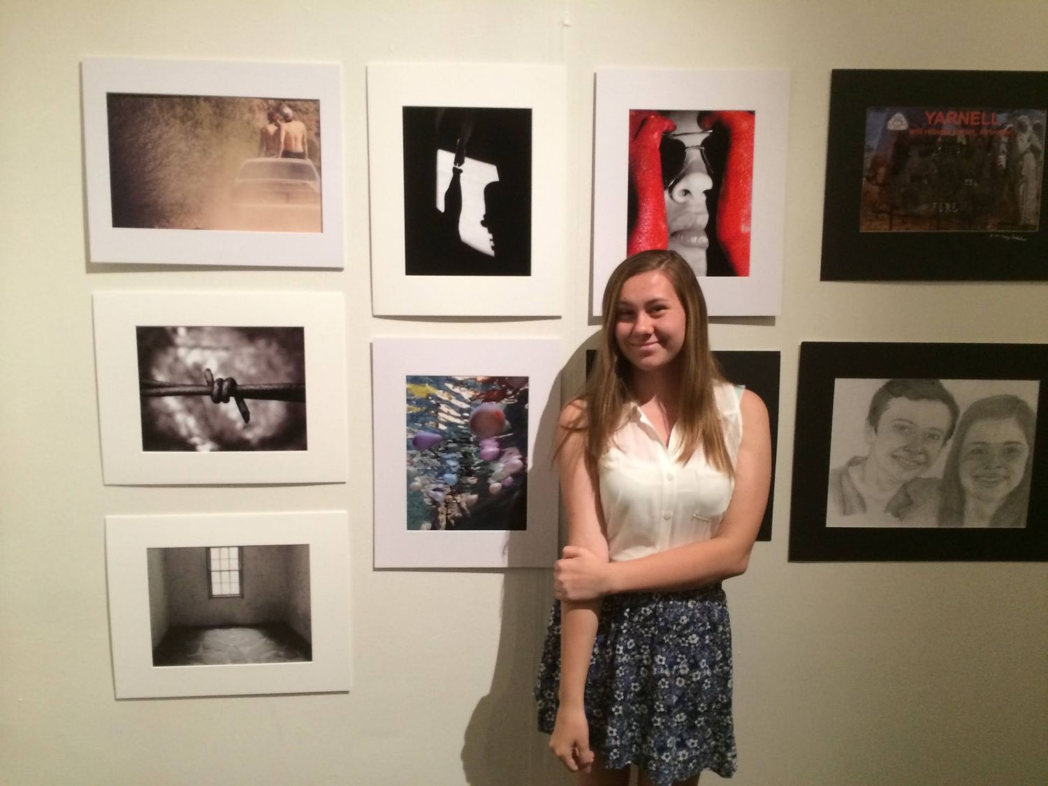 Smiling young woman in a white top and patterned skirt stands beside a gallery wall of framed black-and-white and color student artwork.